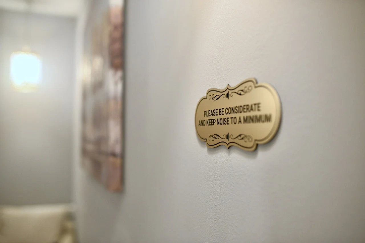 A wall-mounted sign with decorative borders that reads, "Please be considerate and keep noise to a minimum." The sign is attached to a wall with some artwork in the background.