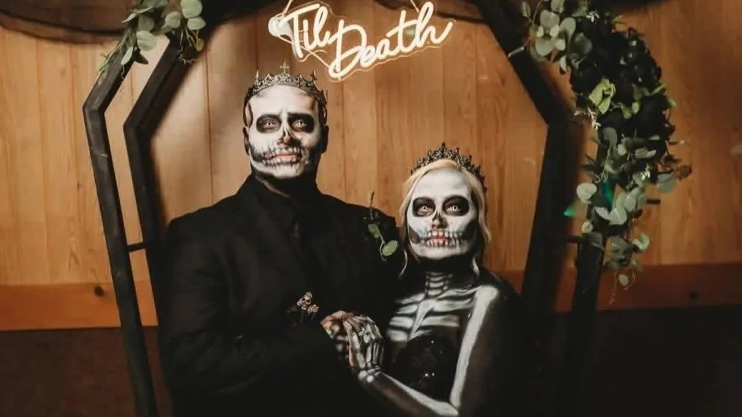 Two people dressed in skeleton and skull makeup with tiaras, posing for a Halloween photo under a sign that says 'Til Death' and surrounded by a wooden frame decorated with green leaves.