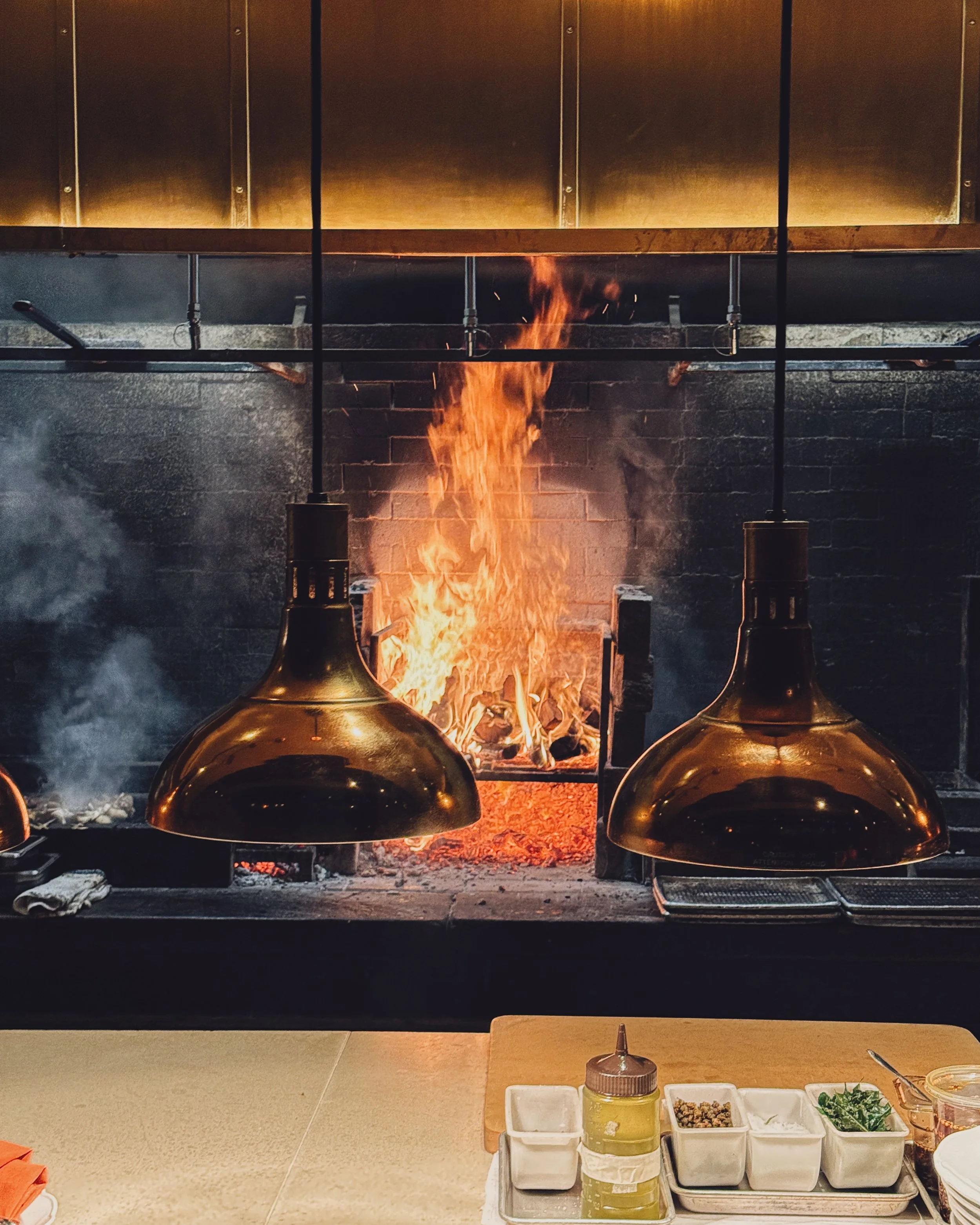 Open kitchen with flames in a brick oven, two black pendant lights hanging over a counter with condiments and herbs.