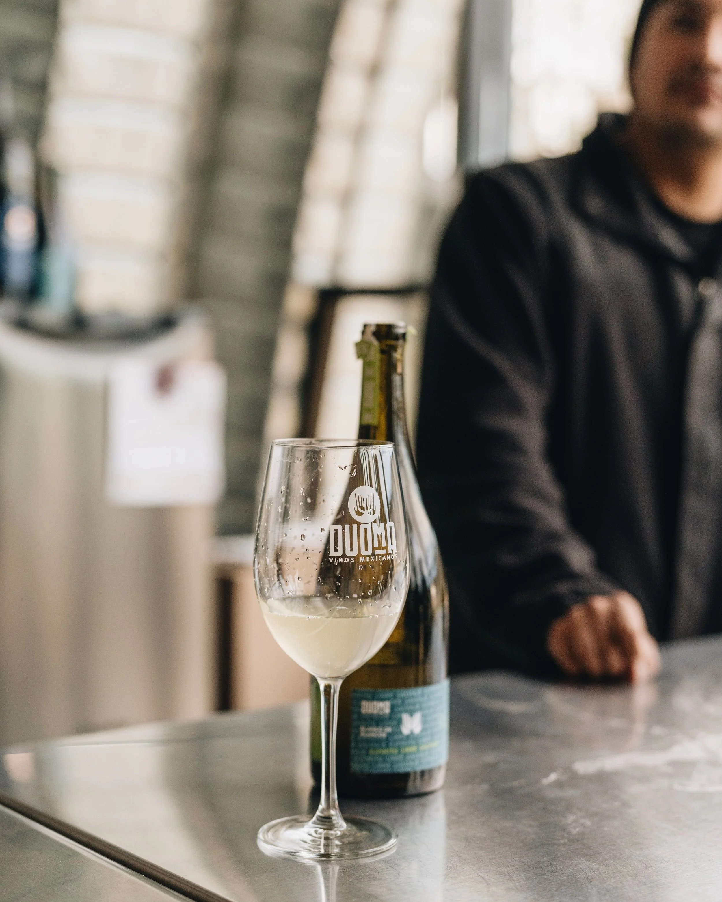 A wine glass with some white liquid inside, placed on a table with a bottle behind it. The glass has a logo that says "Duomo" with a fork, indicating it is from a Mexican winery.