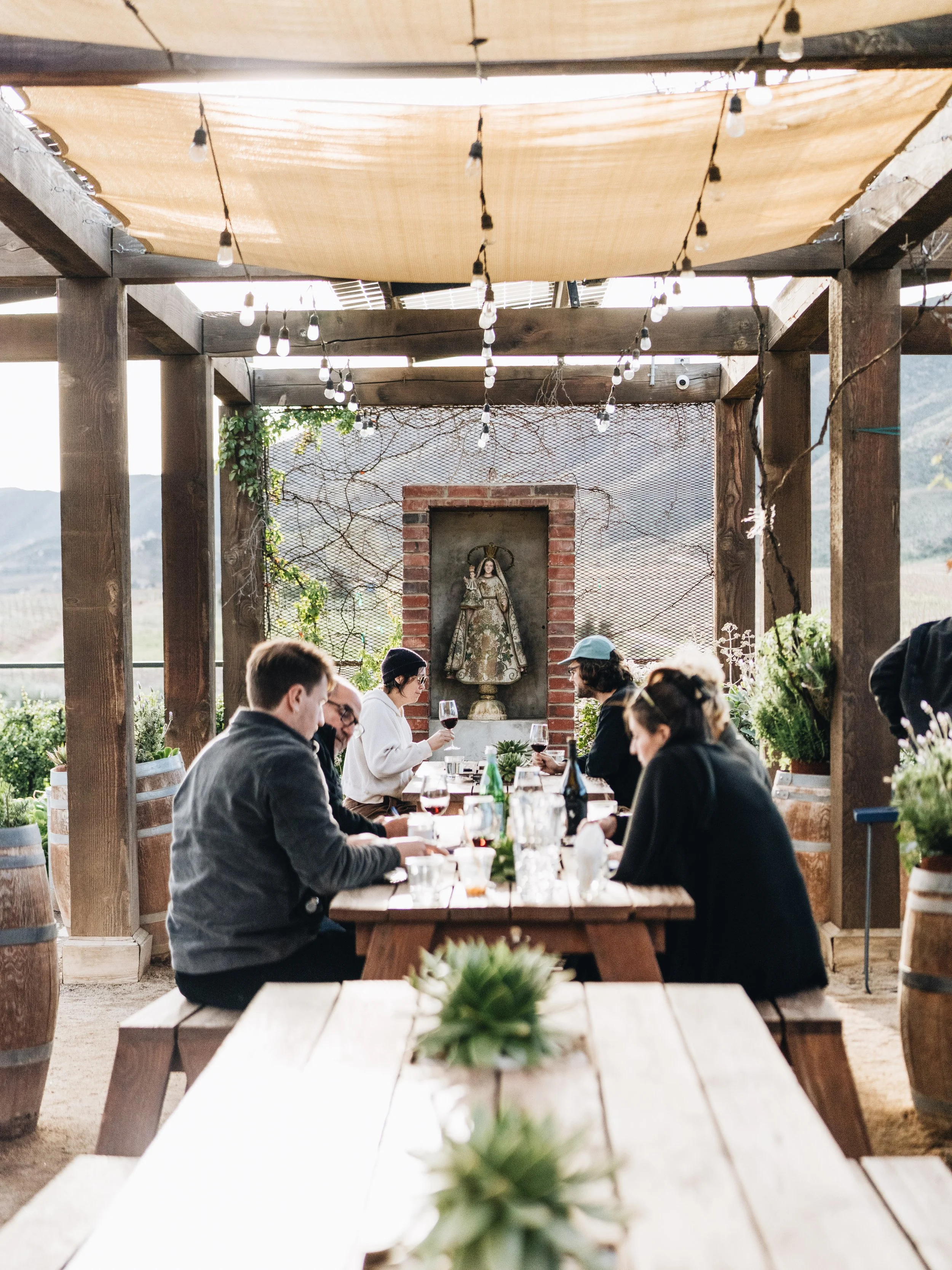 People dining outdoors at a rustic vineyard with a religious statue in the background and string lights overhead.