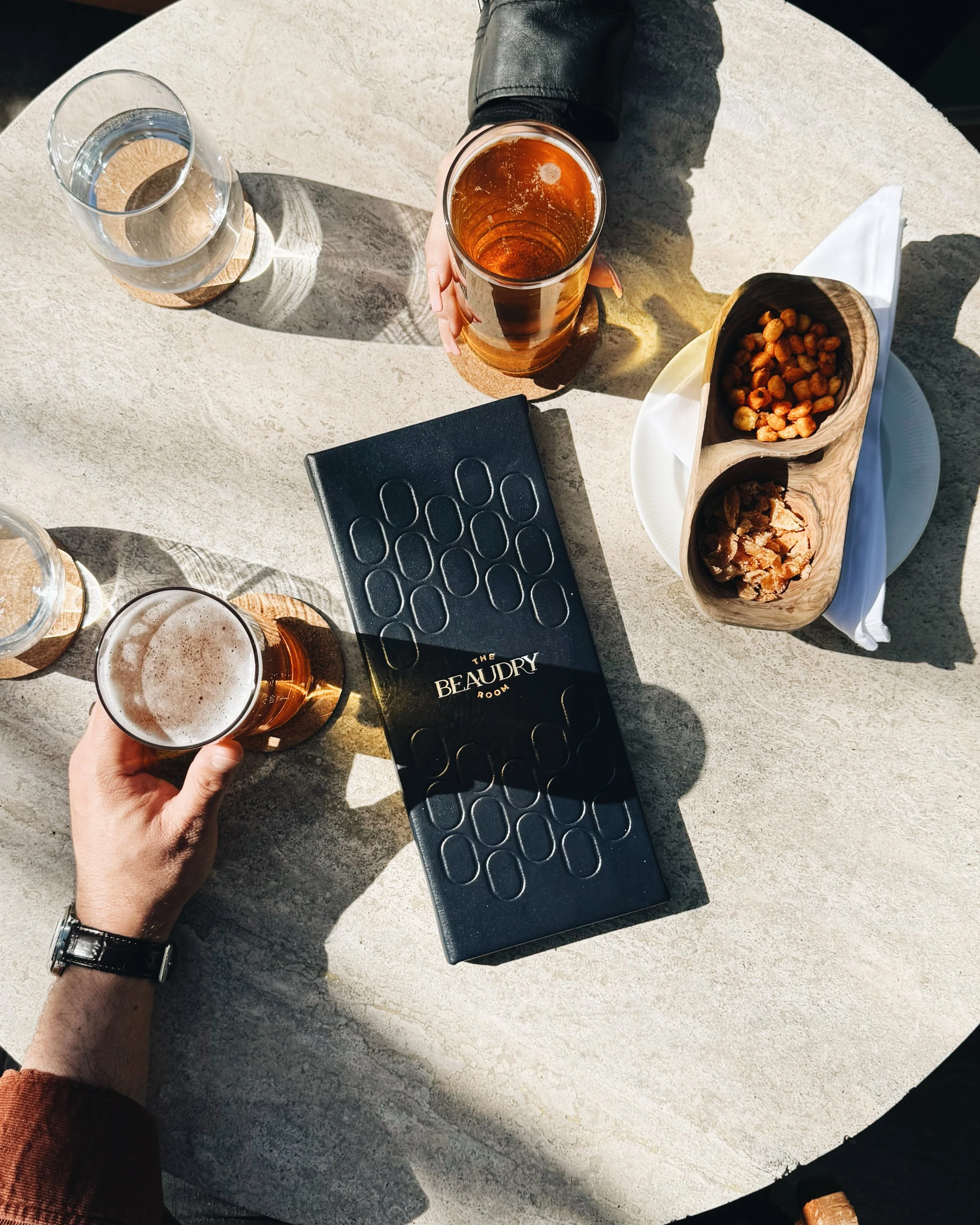 Table with three glasses of beer, a hand holding a glass of beer, a black photo book, and a wooden dish with snacks, including mixed nuts and roasted chickpeas, on a white stone table.