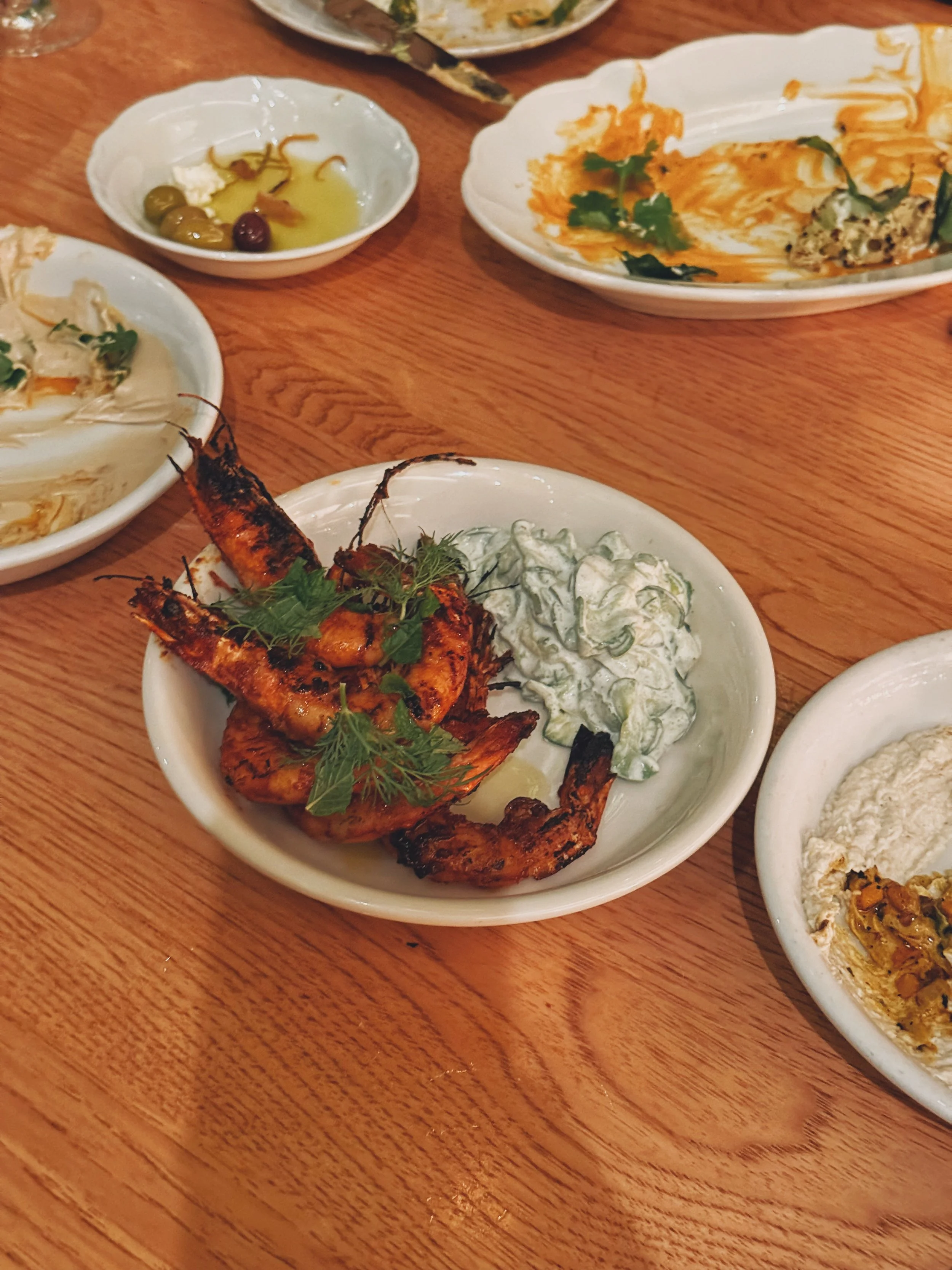 Grilled shrimp with herbs and lemon, accompanied by cucumber salad in a white bowl, served on a wooden table with various dishes and sauces.