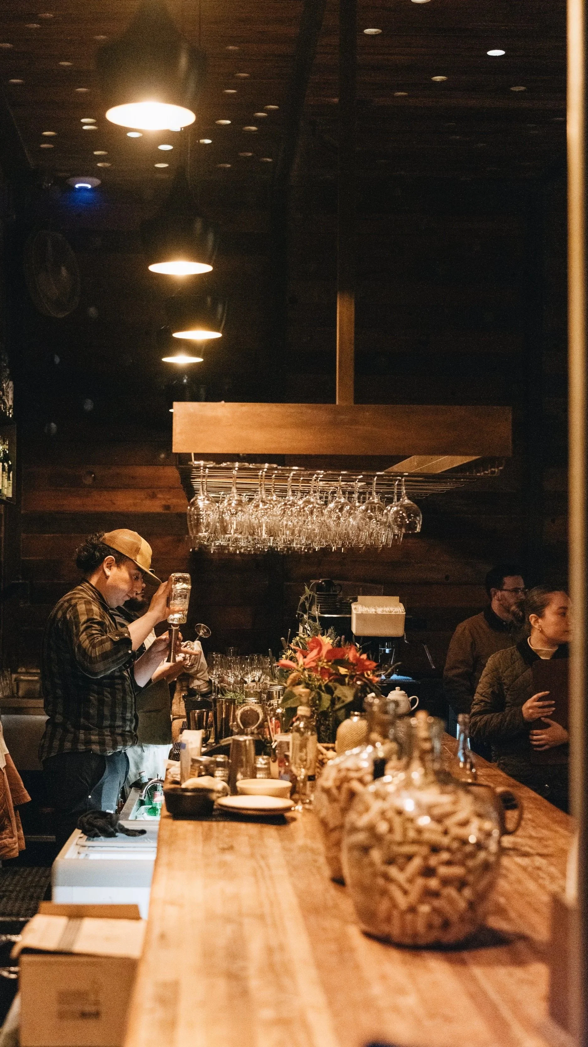 A cozy bar scene with a bartender preparing drinks, glasses hanging above, and customers waiting nearby in a warmly lit wooden interior decorated with flowers.