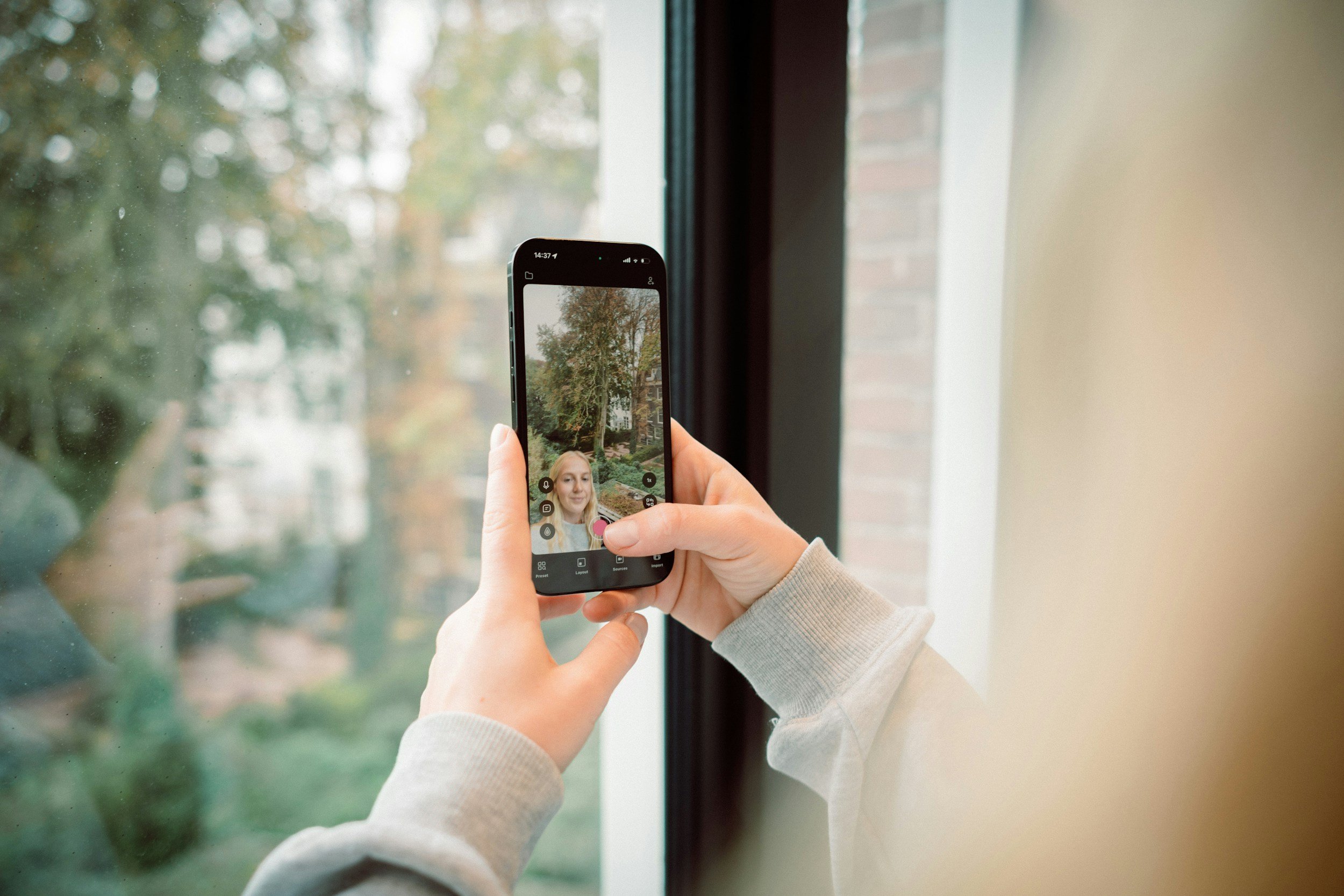 Person taking a photo of a woman through a window using a smartphone.