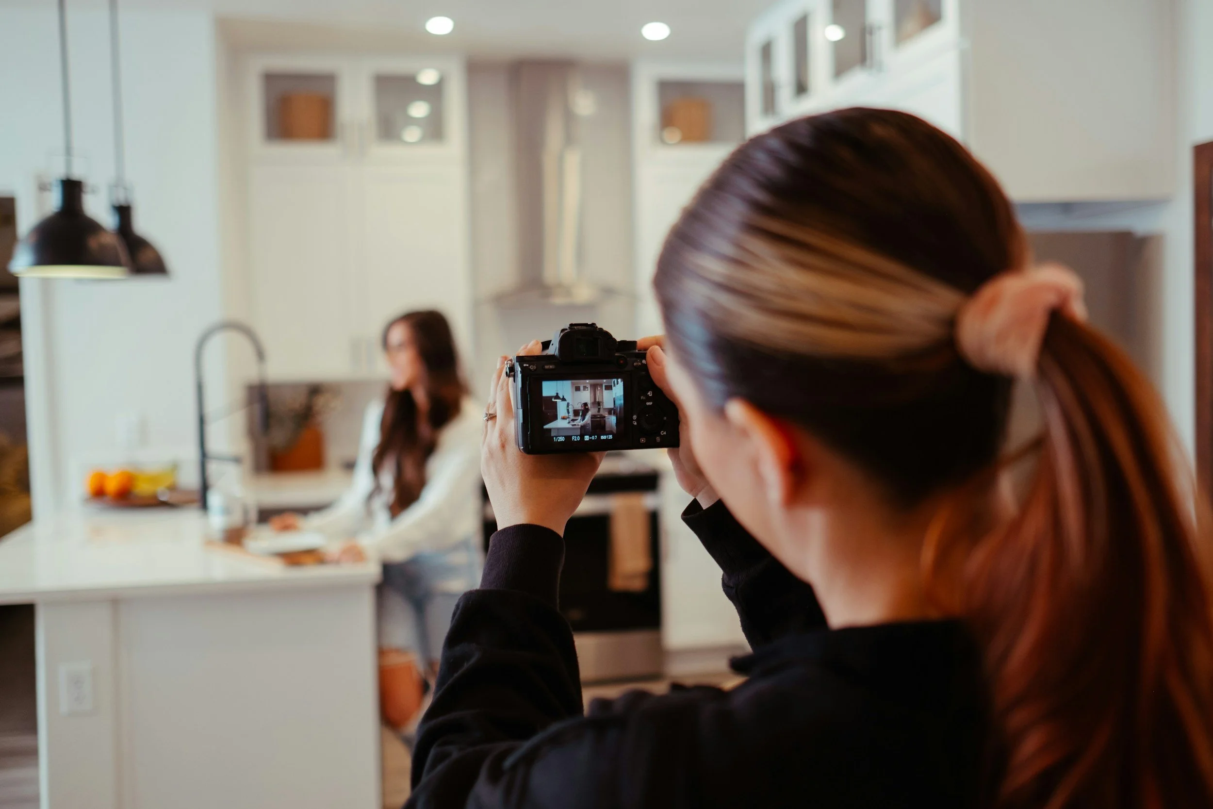 A woman taking a photo of another woman seated at a kitchen counter with a camera.