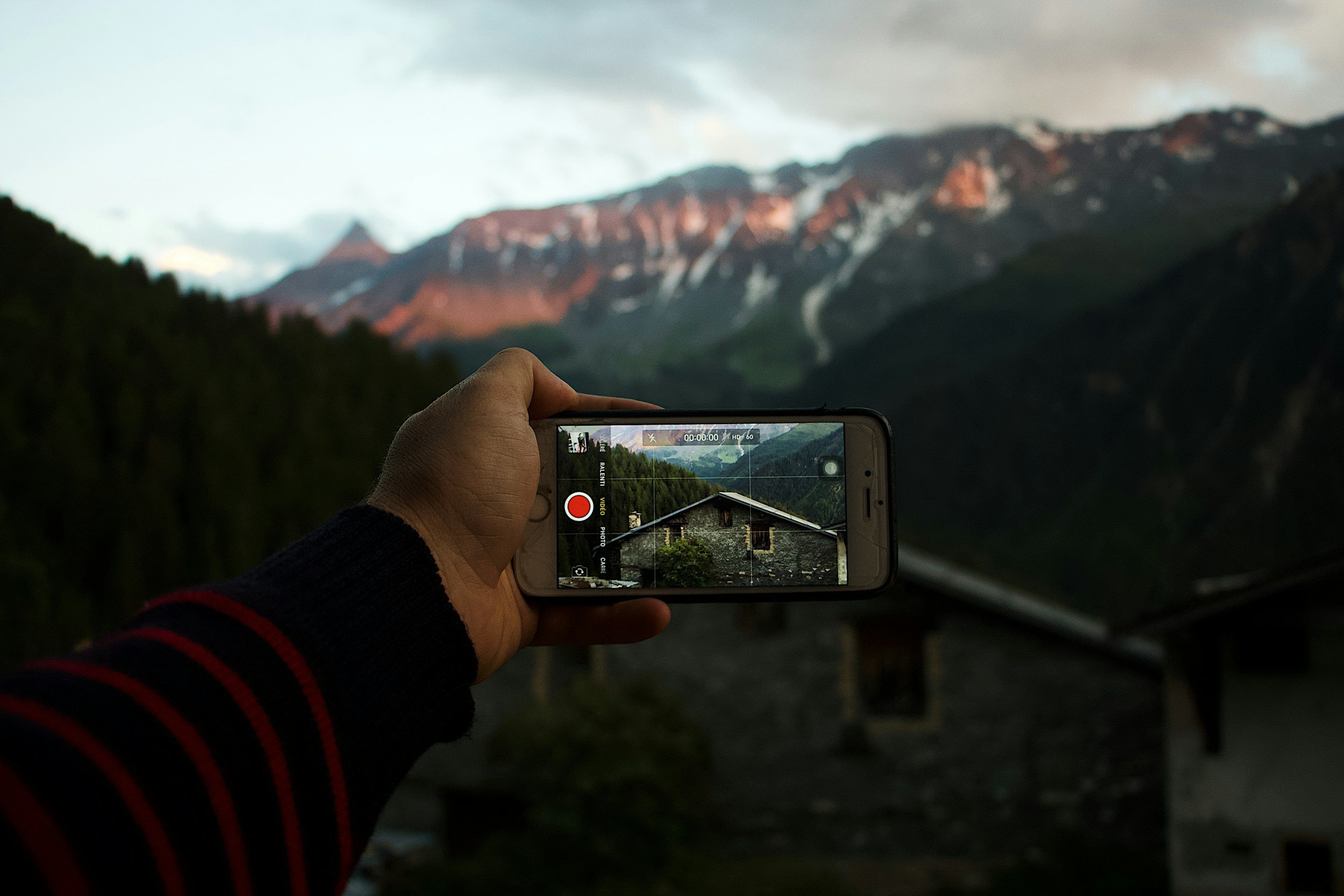 Person holding a smartphone taking a photo of a mountain landscape with a house in the foreground.
