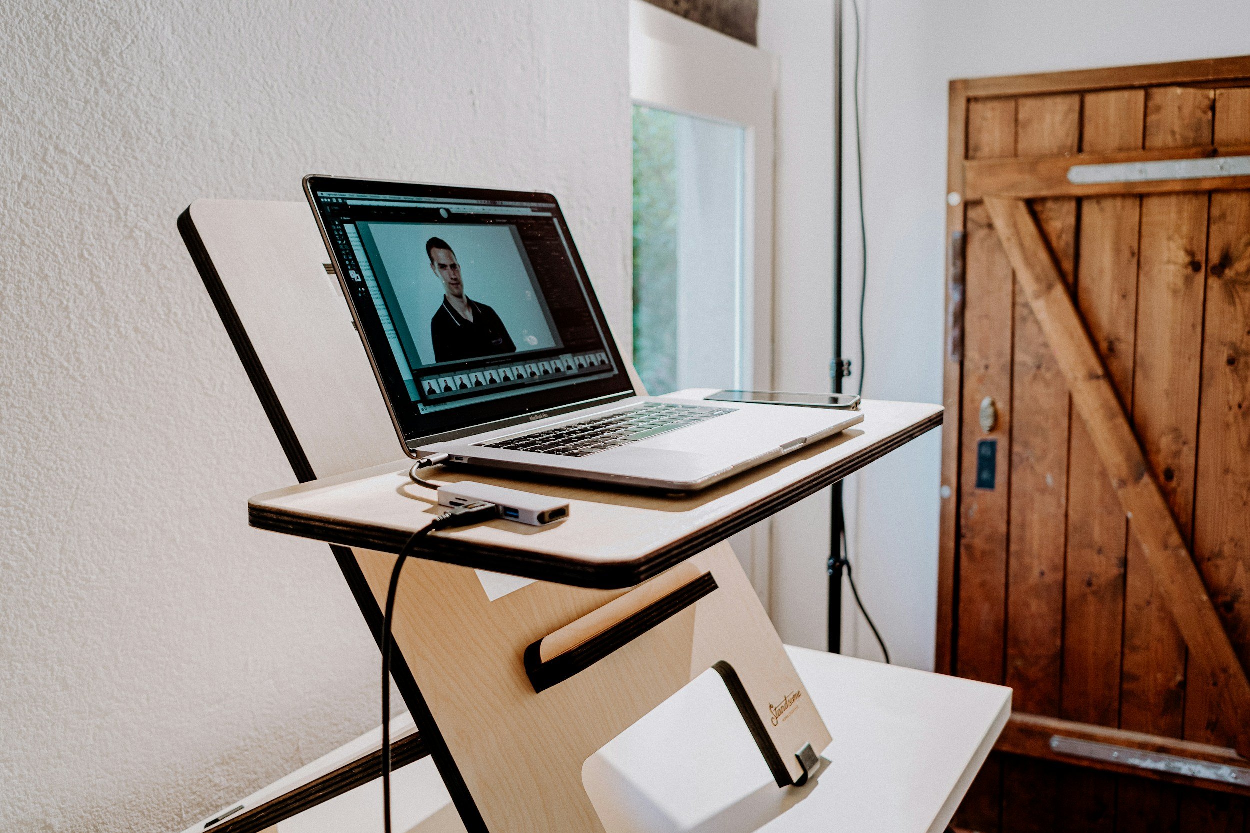 Laptop on a stand showing a photo editing software with a portrait of a man, placed on a small table near a white wall and a wooden door.