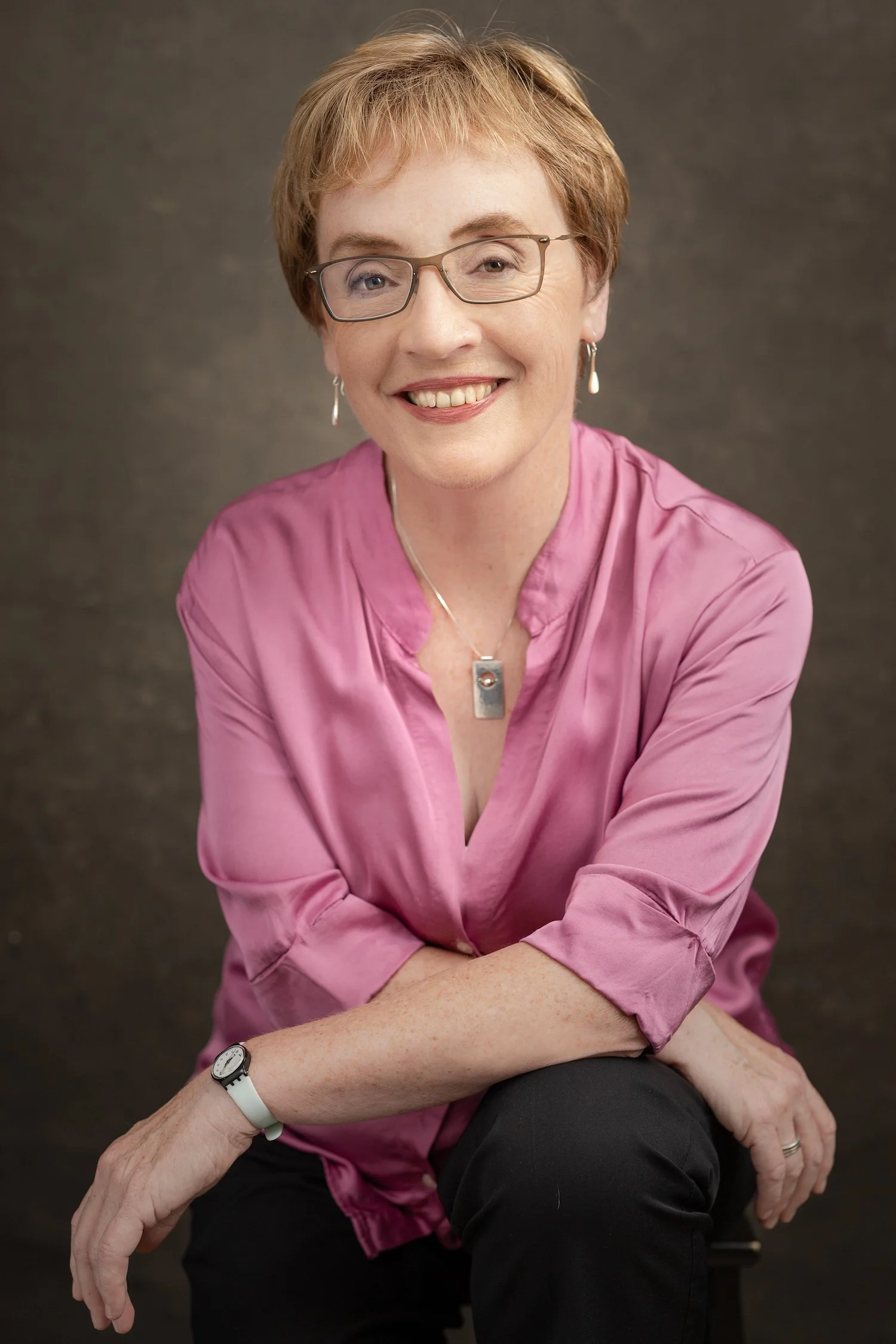 A smiling woman with short red hair wearing glasses, a pink satin blouse, and black pants, sitting with her arms crossed, against a dark background.