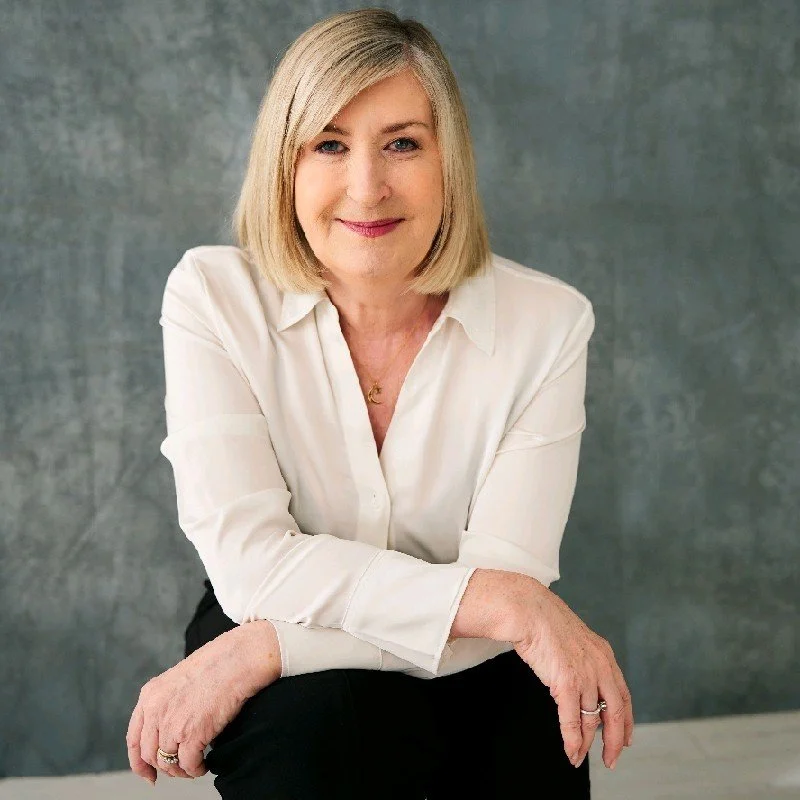 A woman with blonde hair wearing a white button-down shirt, sitting with one arm resting on her knee, smiling at the camera, in front of a gray background.