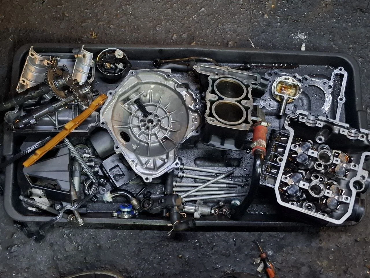Disassembled car engine parts laid out on a black tray, including pistons, cylinder head, valves, and various metal components.