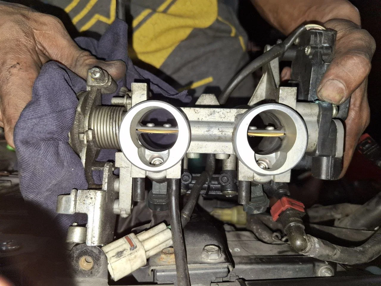 A person wearing gloves working on a carburetor, holding it with both hands. The carburetor is metallic with visible parts, including the throttle plates and linkages.