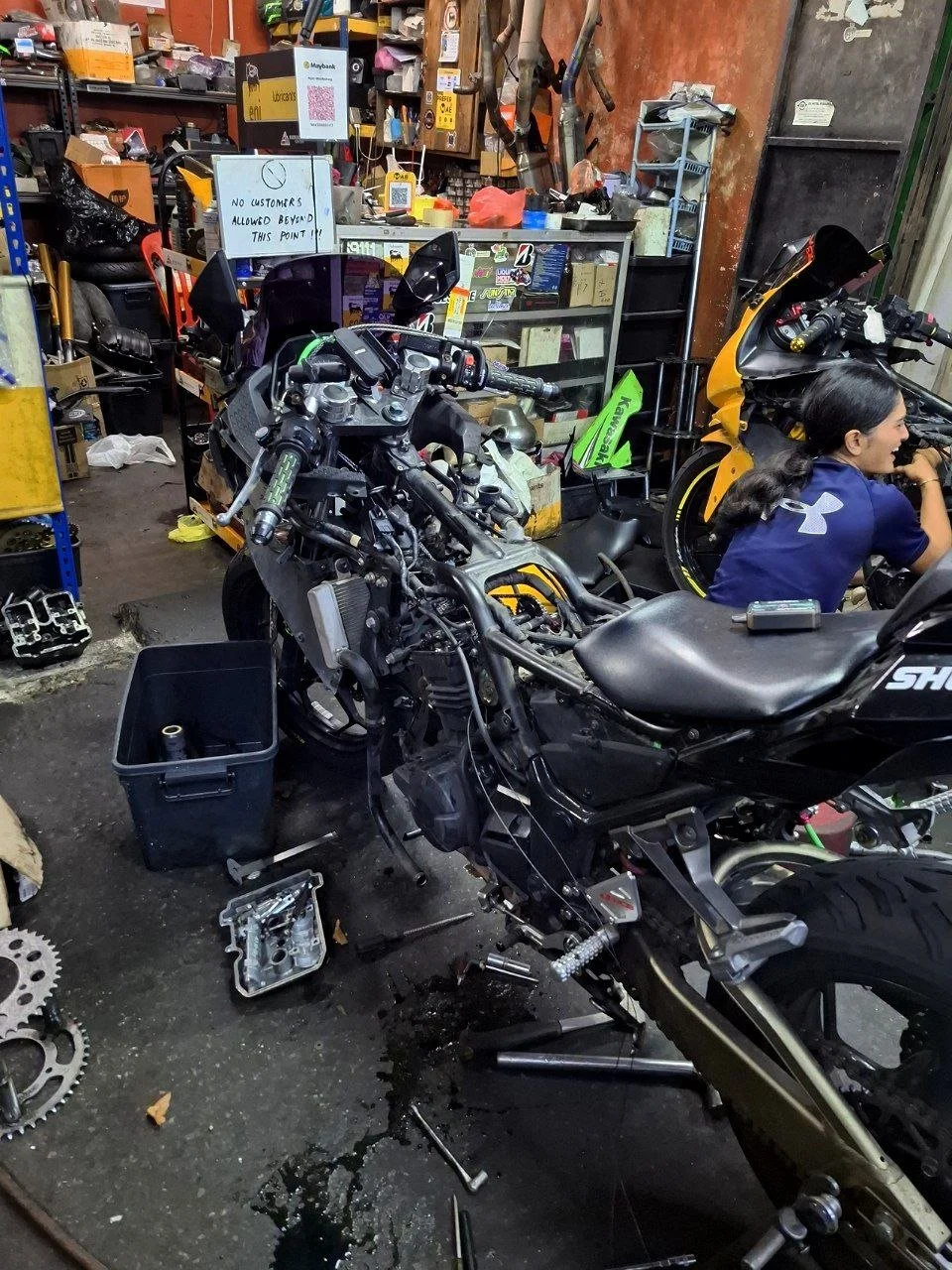 A motorcycle in a workshop undergoing repair, with parts, tools, and a woman working on another motorcycle nearby.
