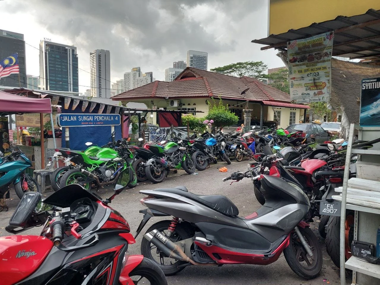 Parking area filled with motorcycles and scooters in front of a building with a red roof, city skyscrapers in the background, and cloudy sky overhead.
