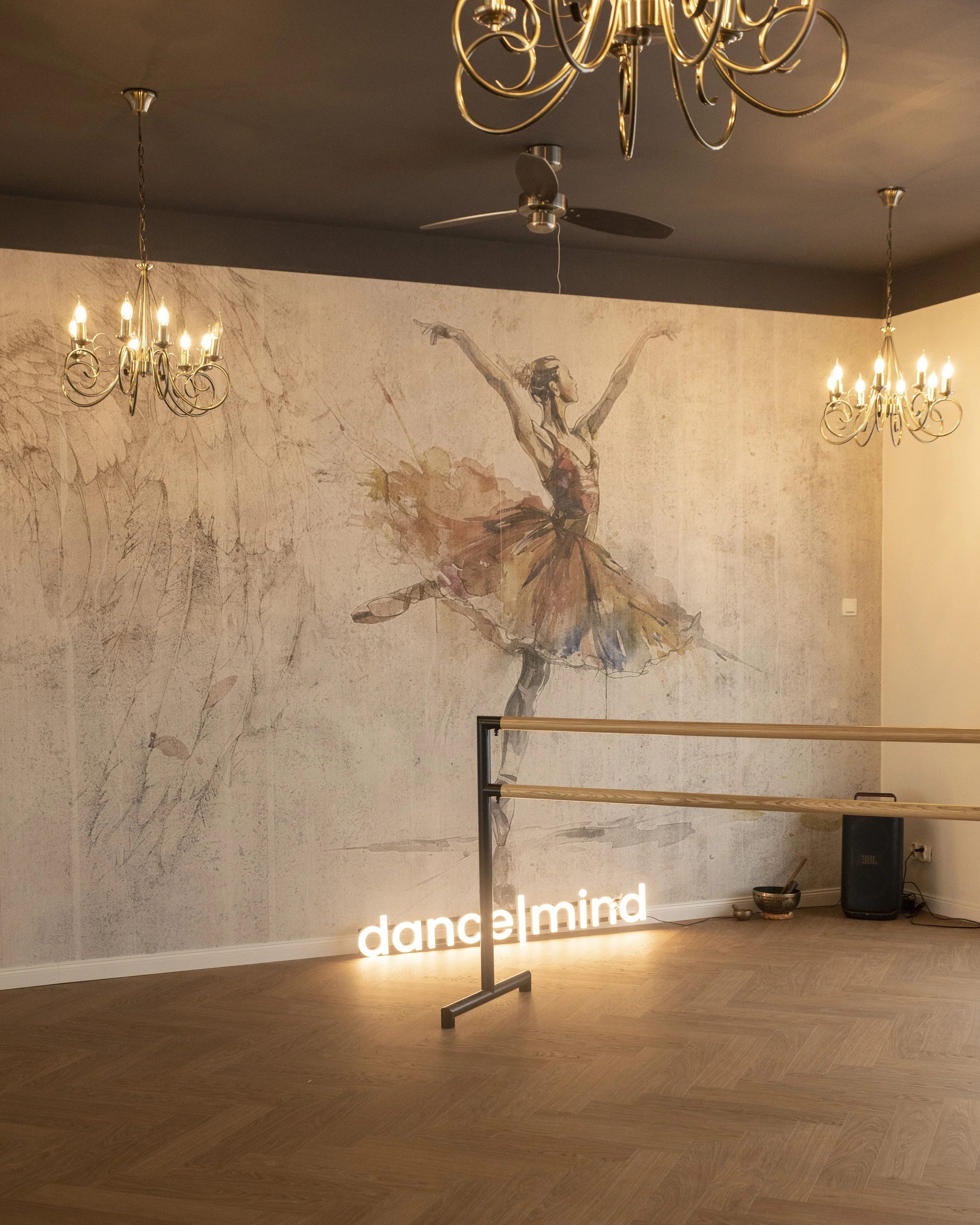 A dance studio with a ballet dancer mural on the wall, chandeliers hanging from the ceiling, wooden flooring, a ballet barre, and a illuminated sign that reads 'dance mind'.