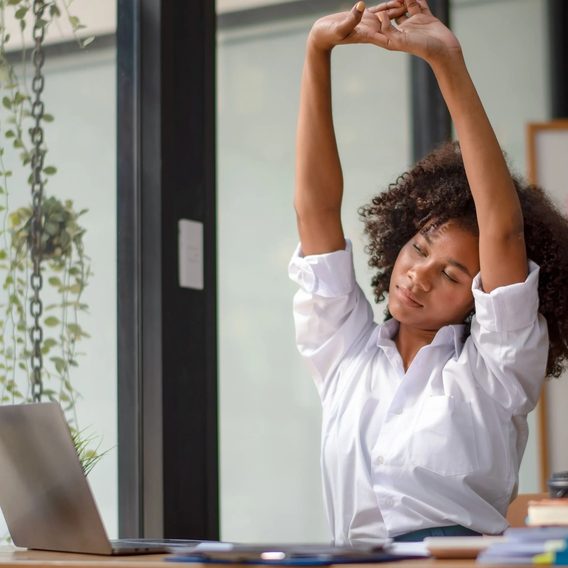 Woman stretching at her desk with a laptop, papers, and plants nearby in a bright office.
