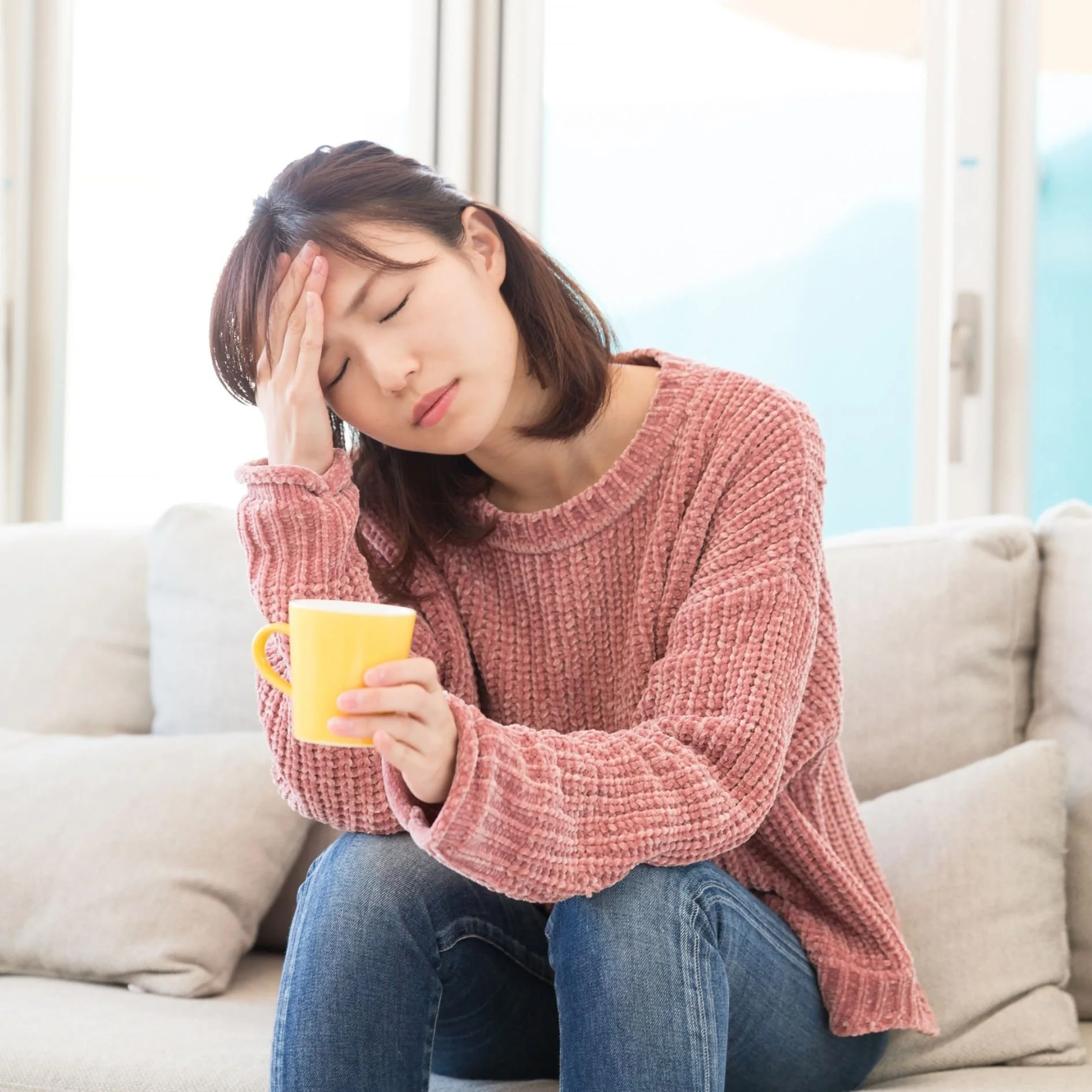A woman sitting on a beige couch holding a yellow coffee mug with her eyes closed and a pained expression on her face, with her hand on her forehead in a bright, casual living room.