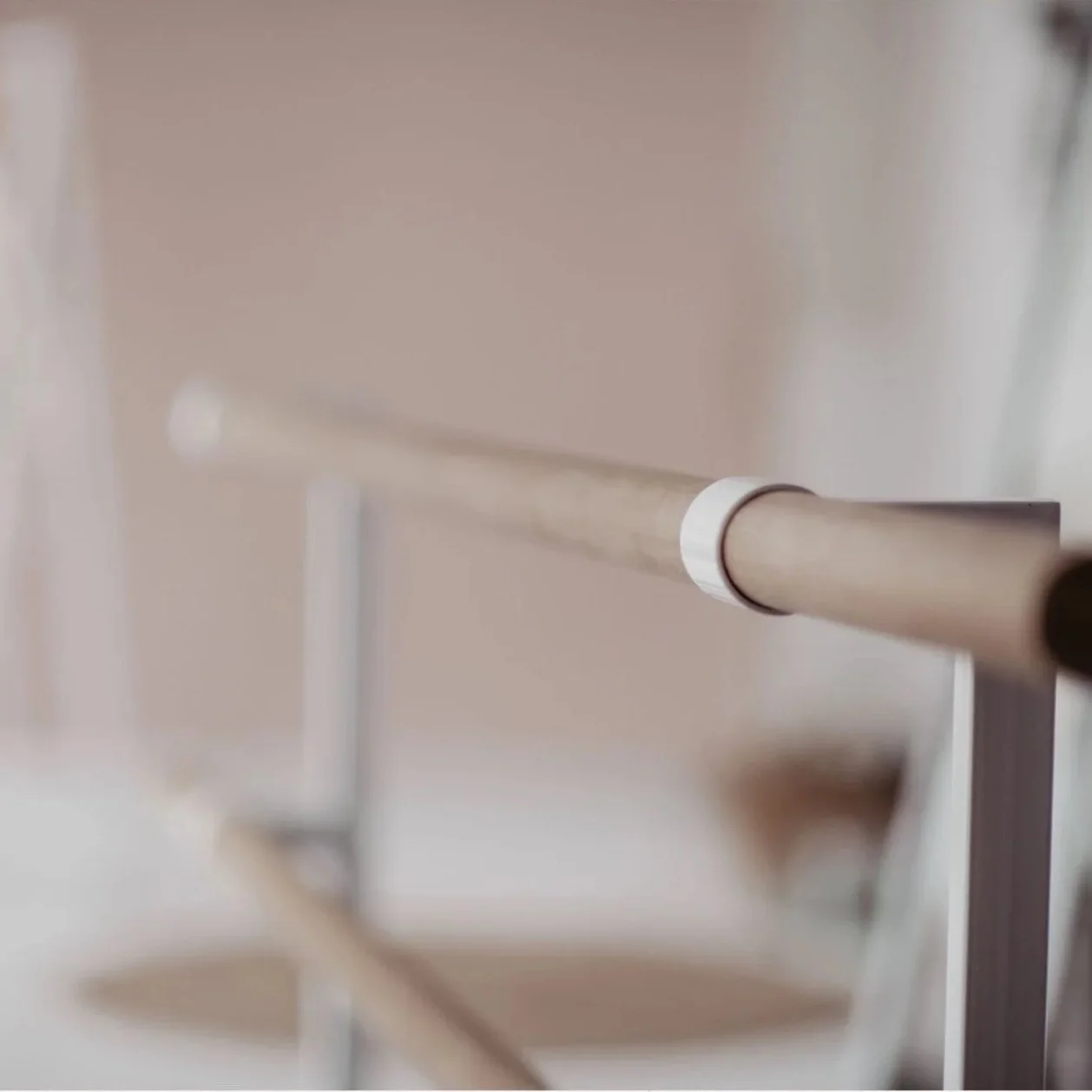 Close-up of a beige or light wooden handrail with a rounded white connector, attached to a metal support, against a plain wall background.