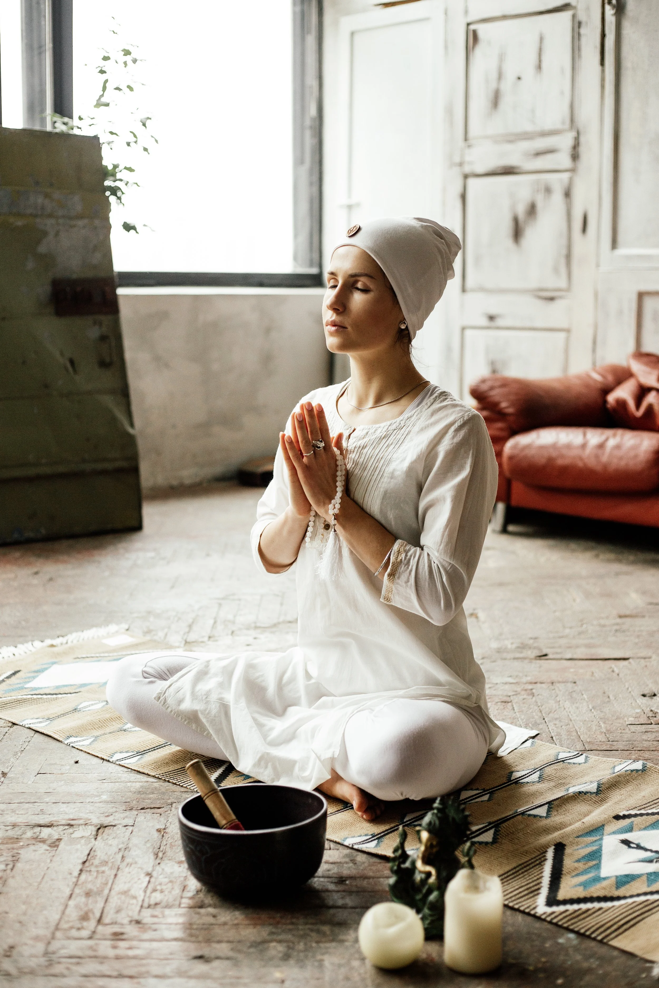 A woman wearing white clothes and a beanie sitting cross-legged on a rug with her hands in prayer position, praying inside a room with rustic decor and candles.