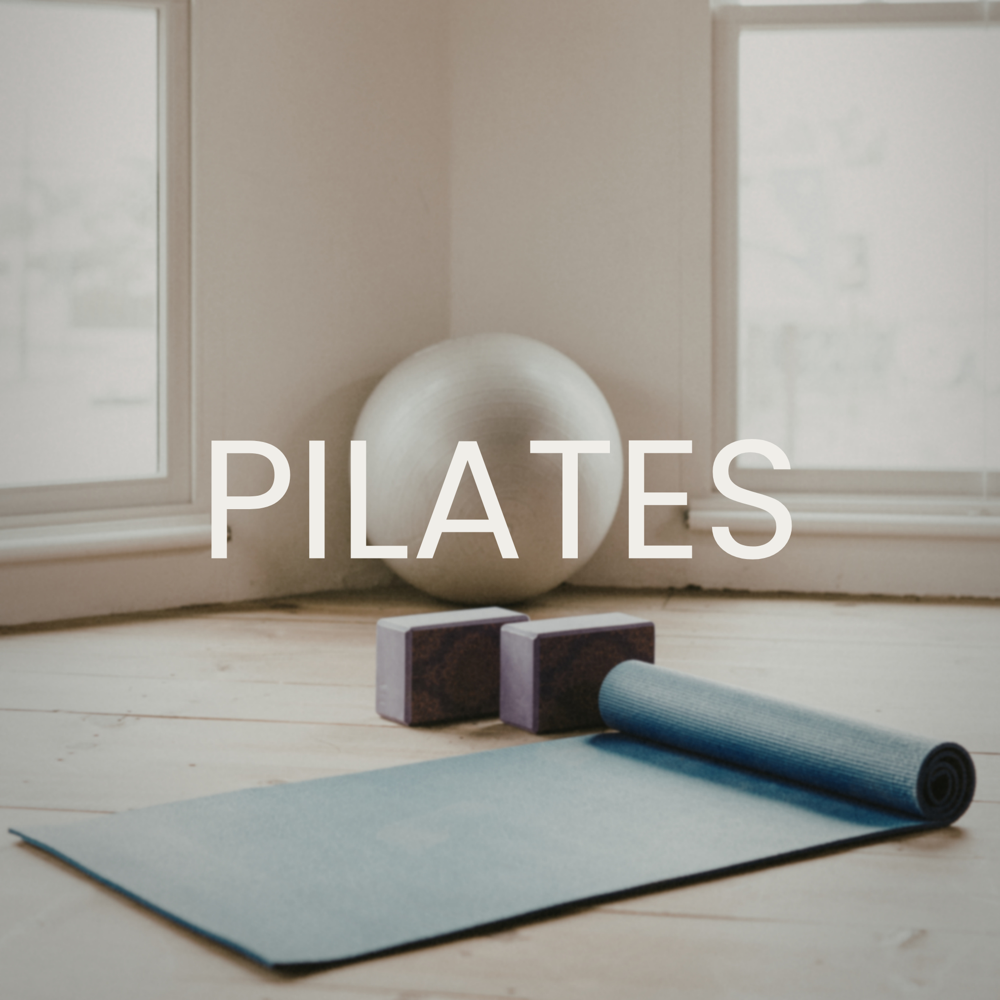 A yoga mat, yoga blocks, and an exercise ball are arranged on a wooden floor near two large windows, with the word 'PILATES' written across the image.
