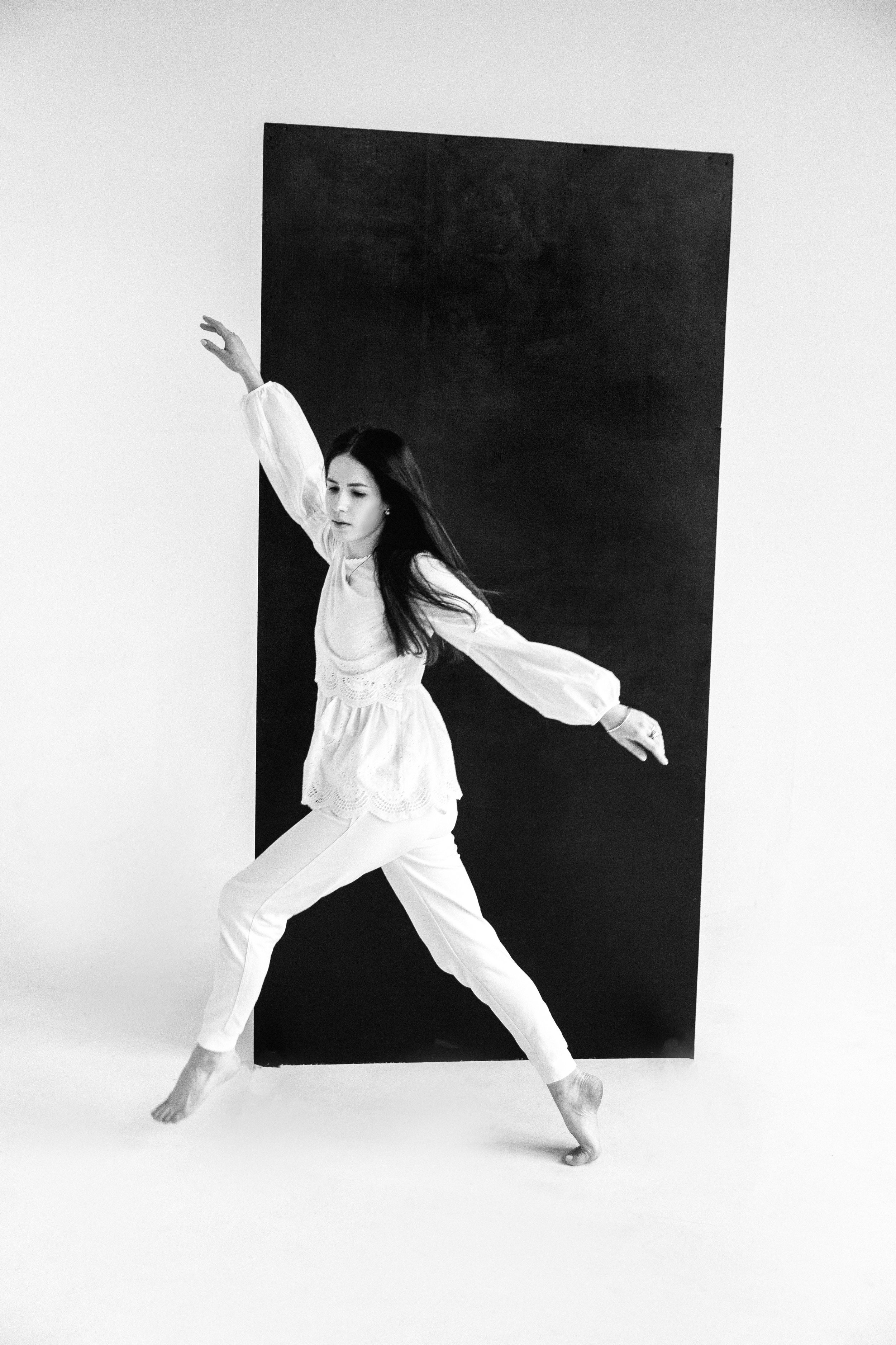 A woman dancing in front of a black panel, dressed in white with long dark hair, captured mid-move on a white studio background.