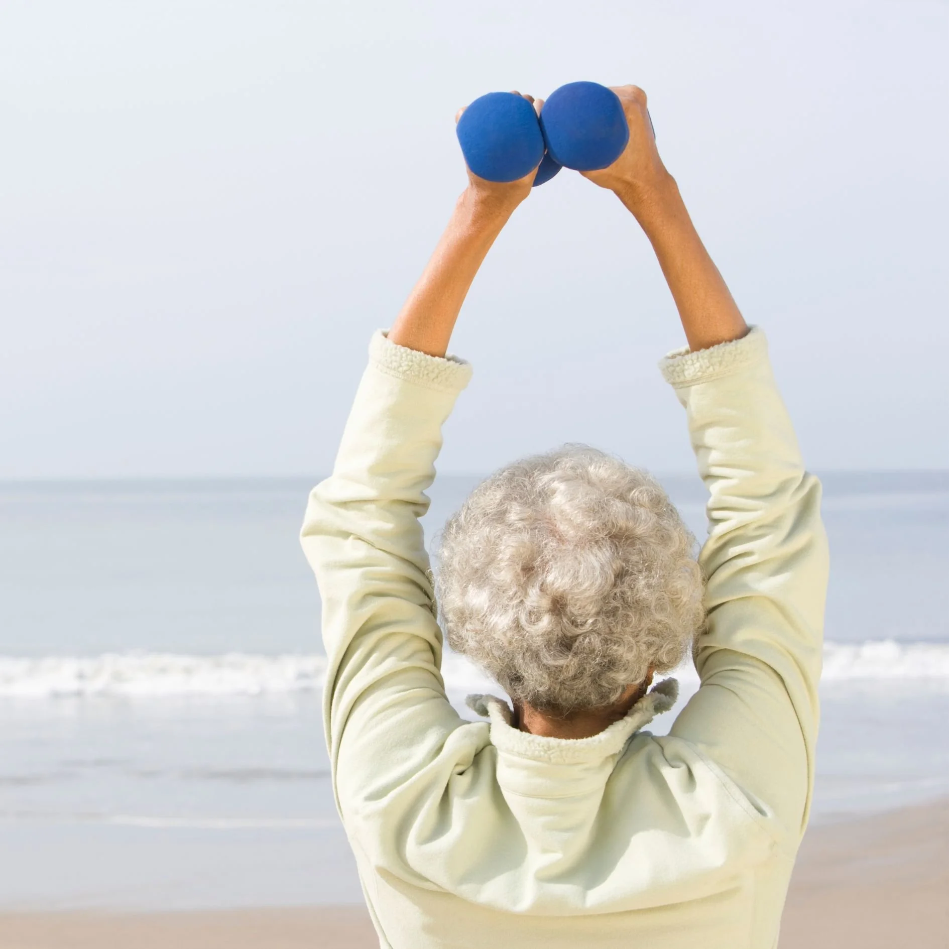Elderly person with white curly hair exercising with small blue dumbbells on a beach.