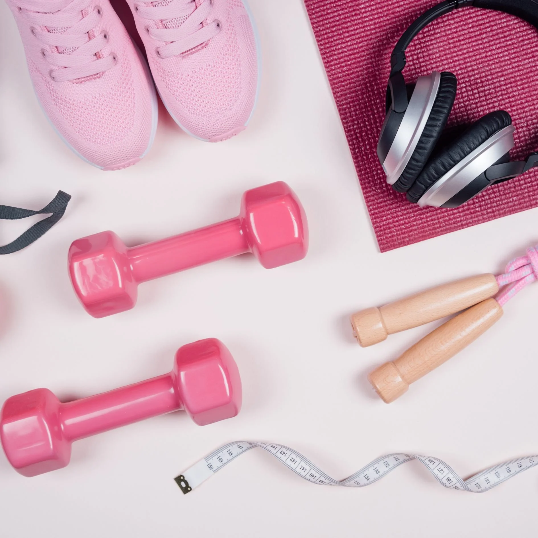Pink sneakers, pink dumbbells, wooden jump rope, headphones on a red workout mat, measuring tape, and pink exercise equipment on a white background.