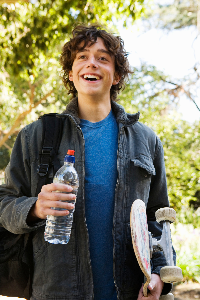 Teen boy smiling and holding a water bottle and skateboard outside