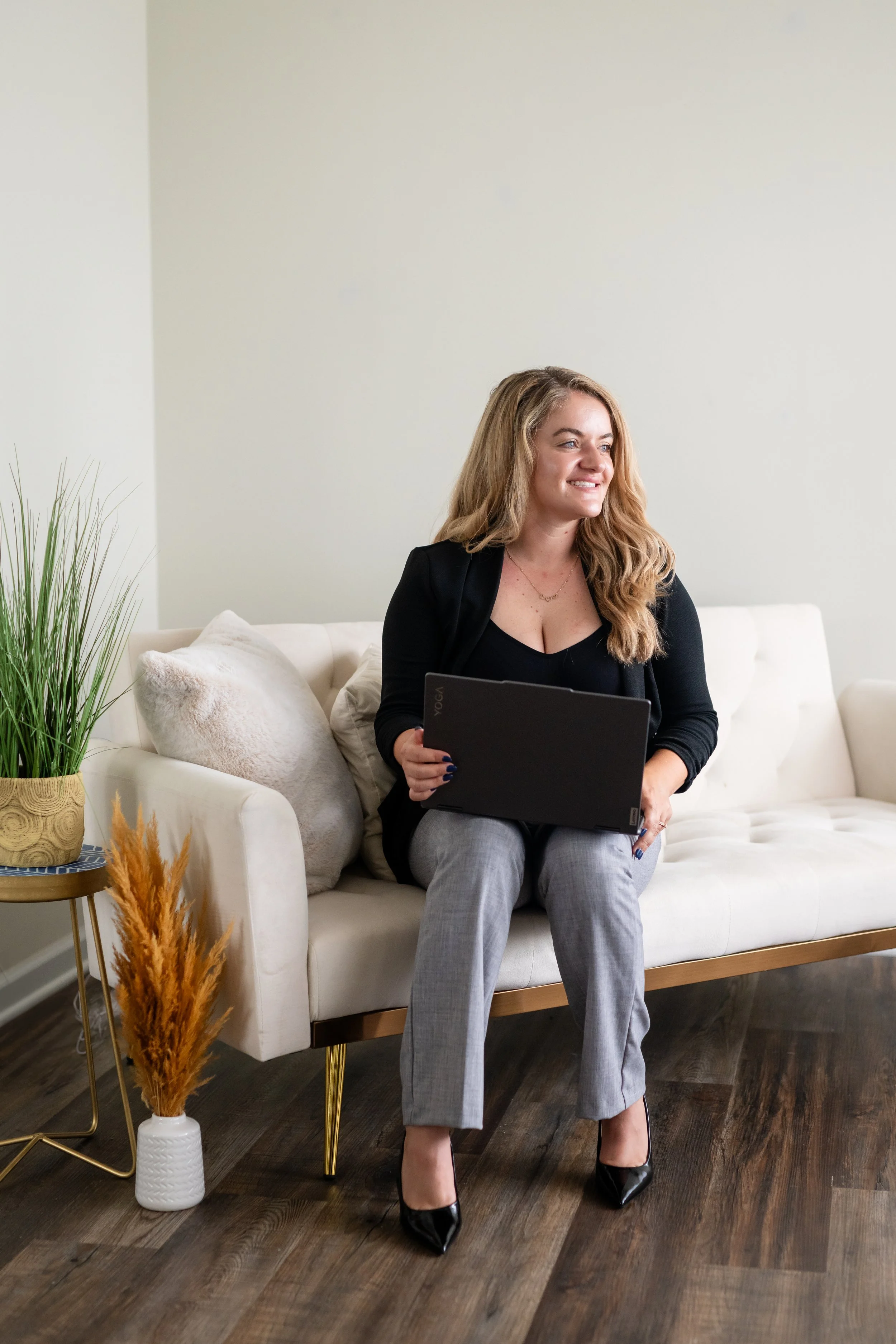 A woman sitting on a white couch holding a black laptop, with a neutral wall background and indoor plants in a decorative pot nearby.