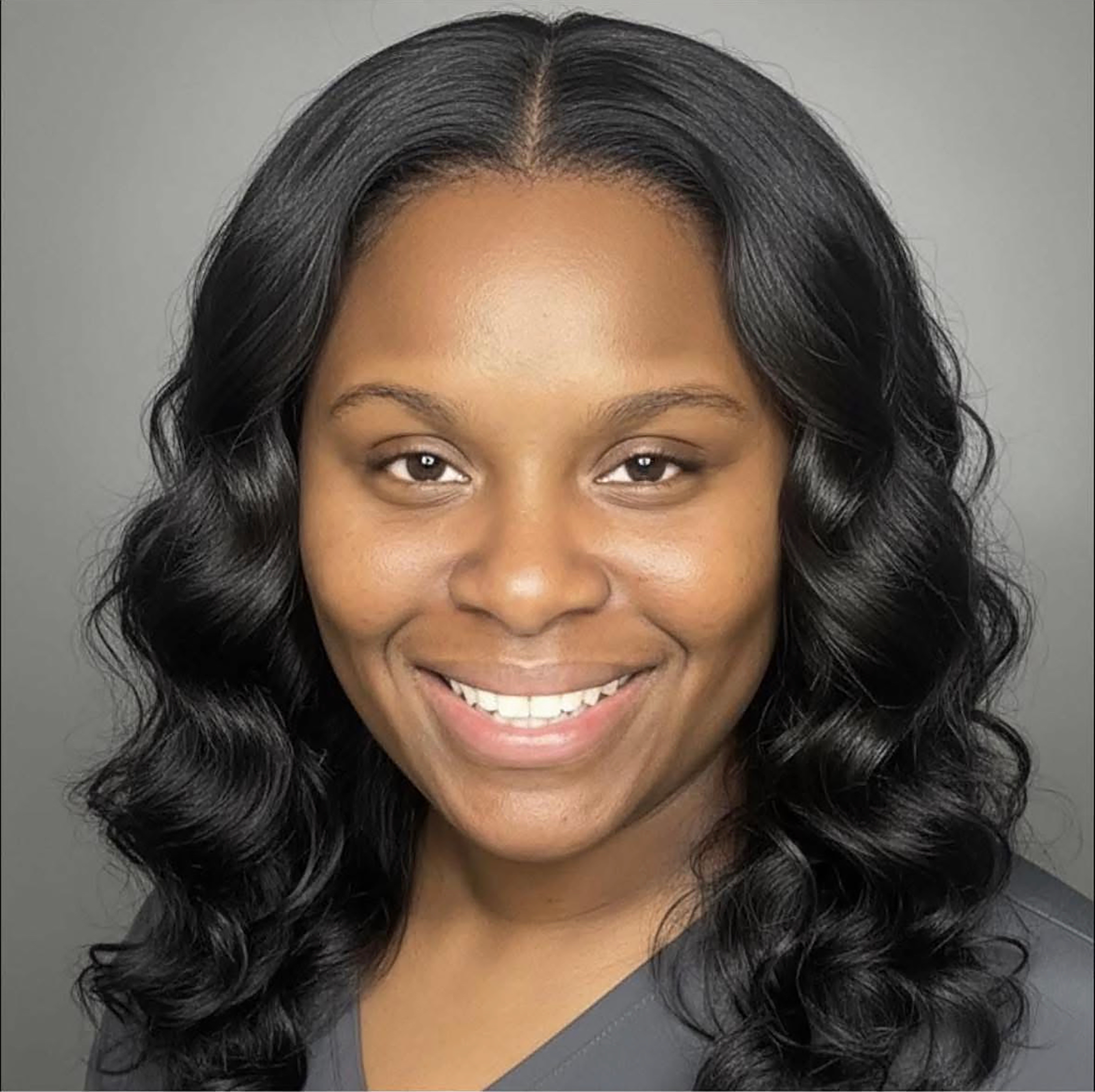 Close-up of a woman with long, curly black hair and a bright smile, wearing a dark gray top, against a plain gray background.