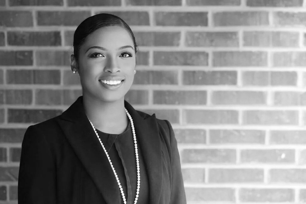 A professional woman with dark hair, smiling, wearing a black blazer, a pearl necklace, and earrings, standing in front of a brick wall.
