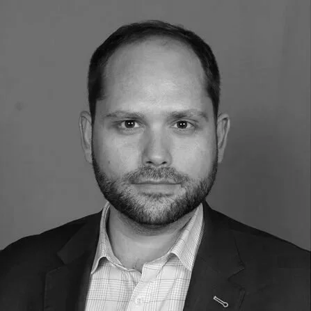 Black and white headshot of a man with short dark hair and a beard, wearing a suit jacket and collared shirt.
