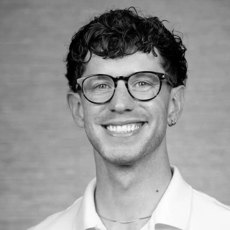 Black and white portrait of a smiling young man with curly hair, glasses, and an earring, wearing a collared shirt, against a plain background.