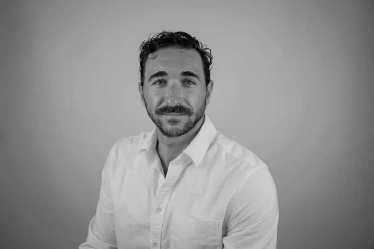 Black and white portrait of a man with dark hair, beard, wearing a white shirt, smiling slightly, against a plain background.