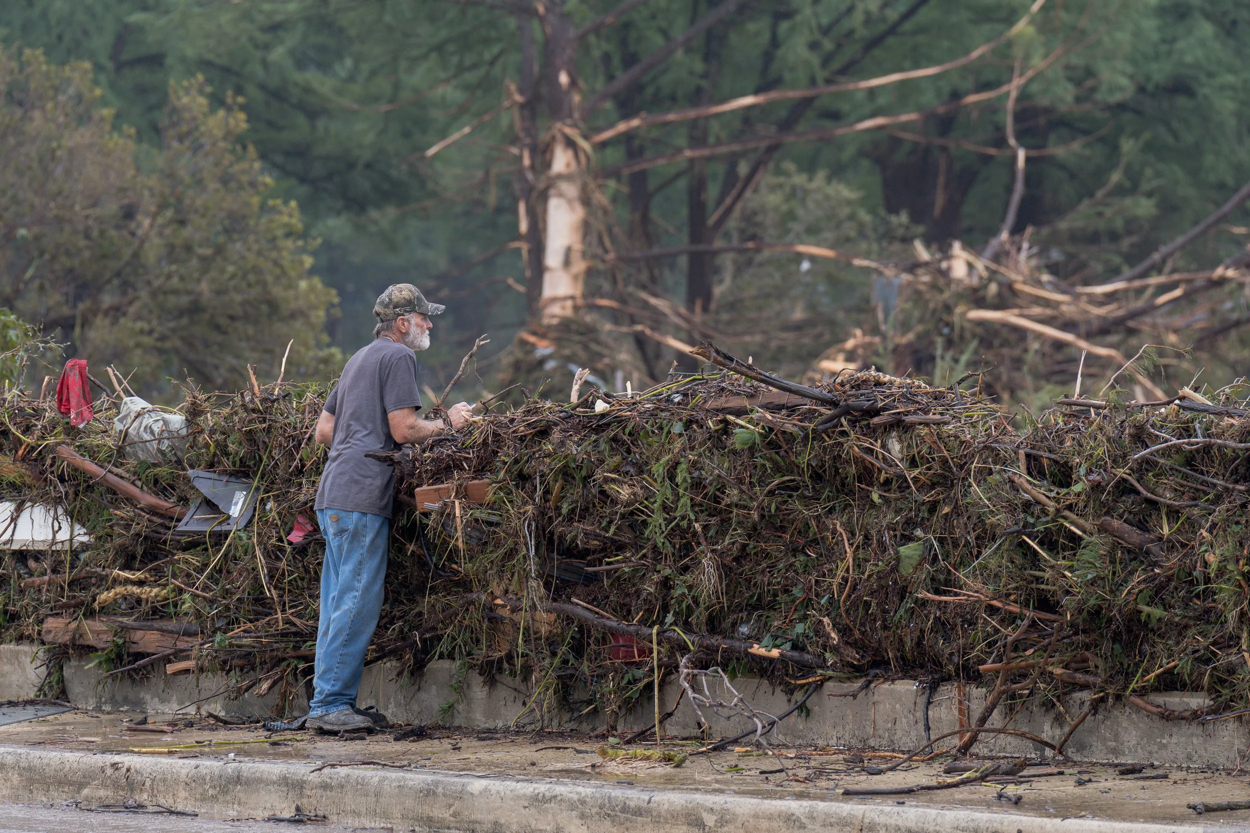 A man in a gray T-shirt and camo cap examines a huge tangle of flood debris stacked along a bridge rail after the Guadalupe River overflowed its banks.