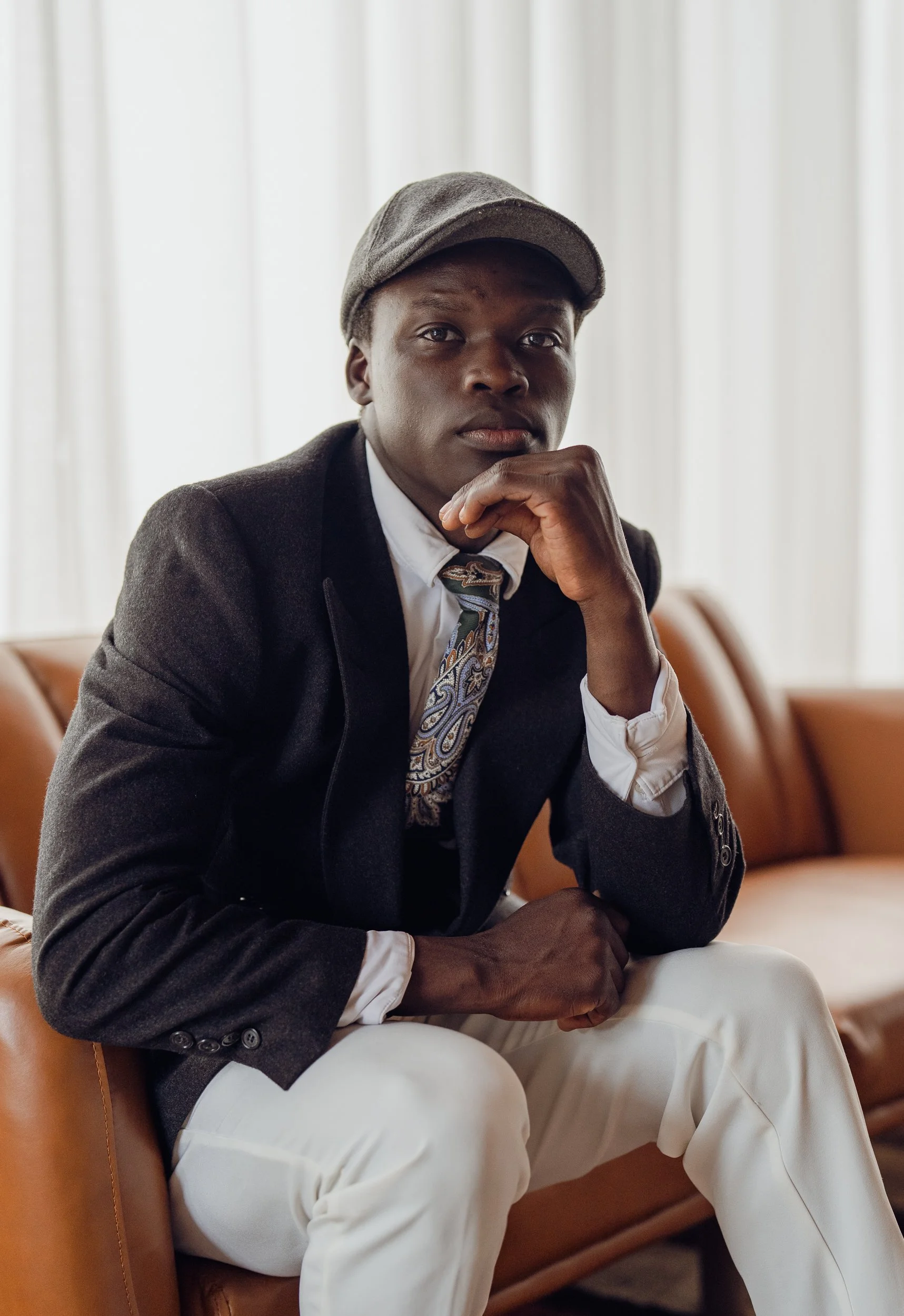 A young man in a gray cap, dark blazer and paisley tie sits on a brown leather sofa, resting his chin on his hand and looking at the camera.