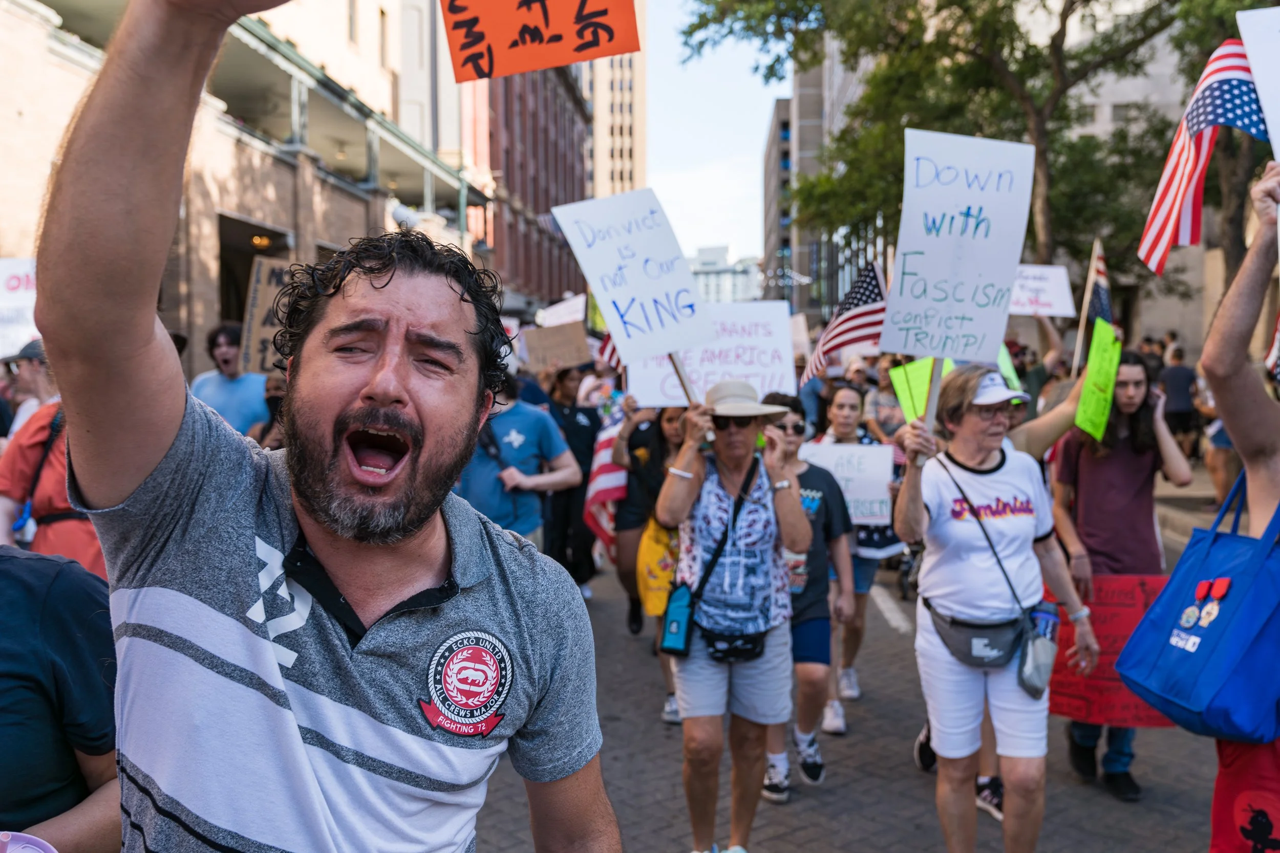A man with a beard and curly hair shouting at a protest march, surrounded by people holding signs and American flags on a city street.