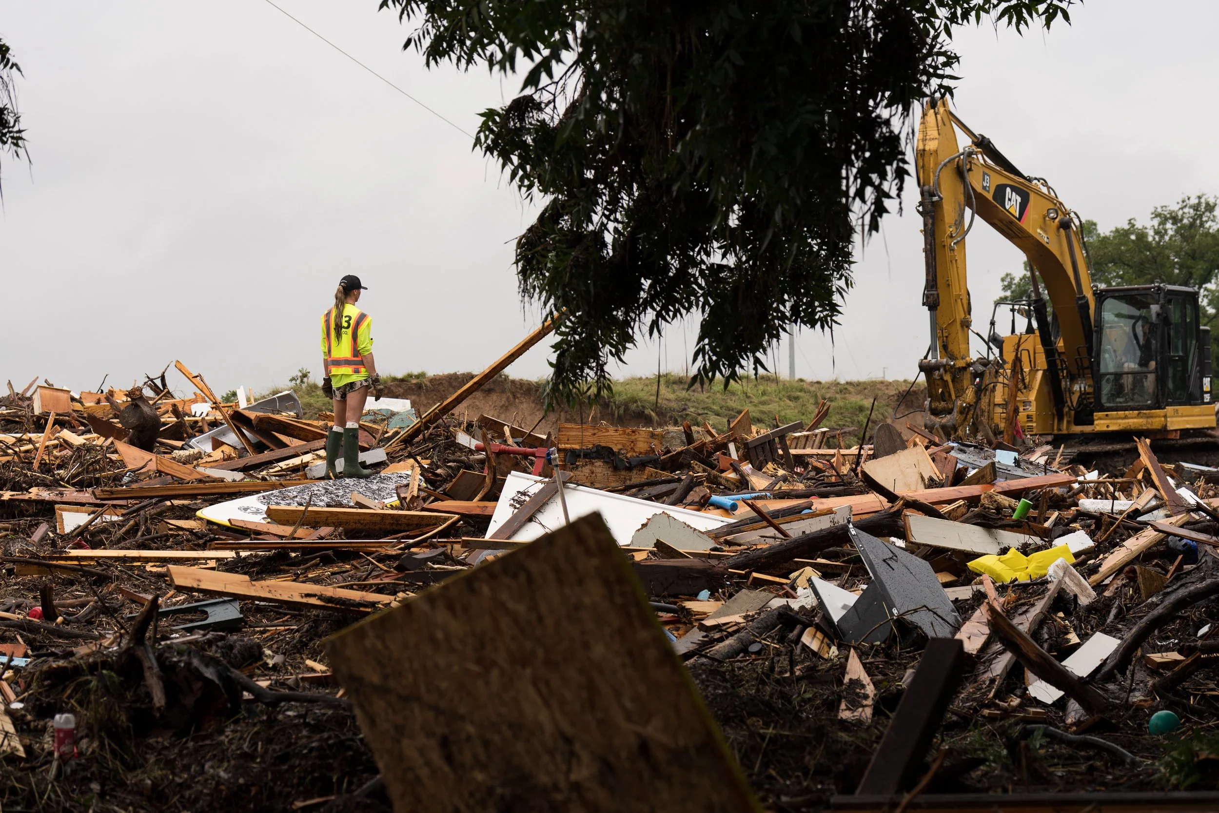 A worker in a yellow safety vest stands amid broken lumber and metal as an excavator clears flood-damaged structures along the Guadalupe River.