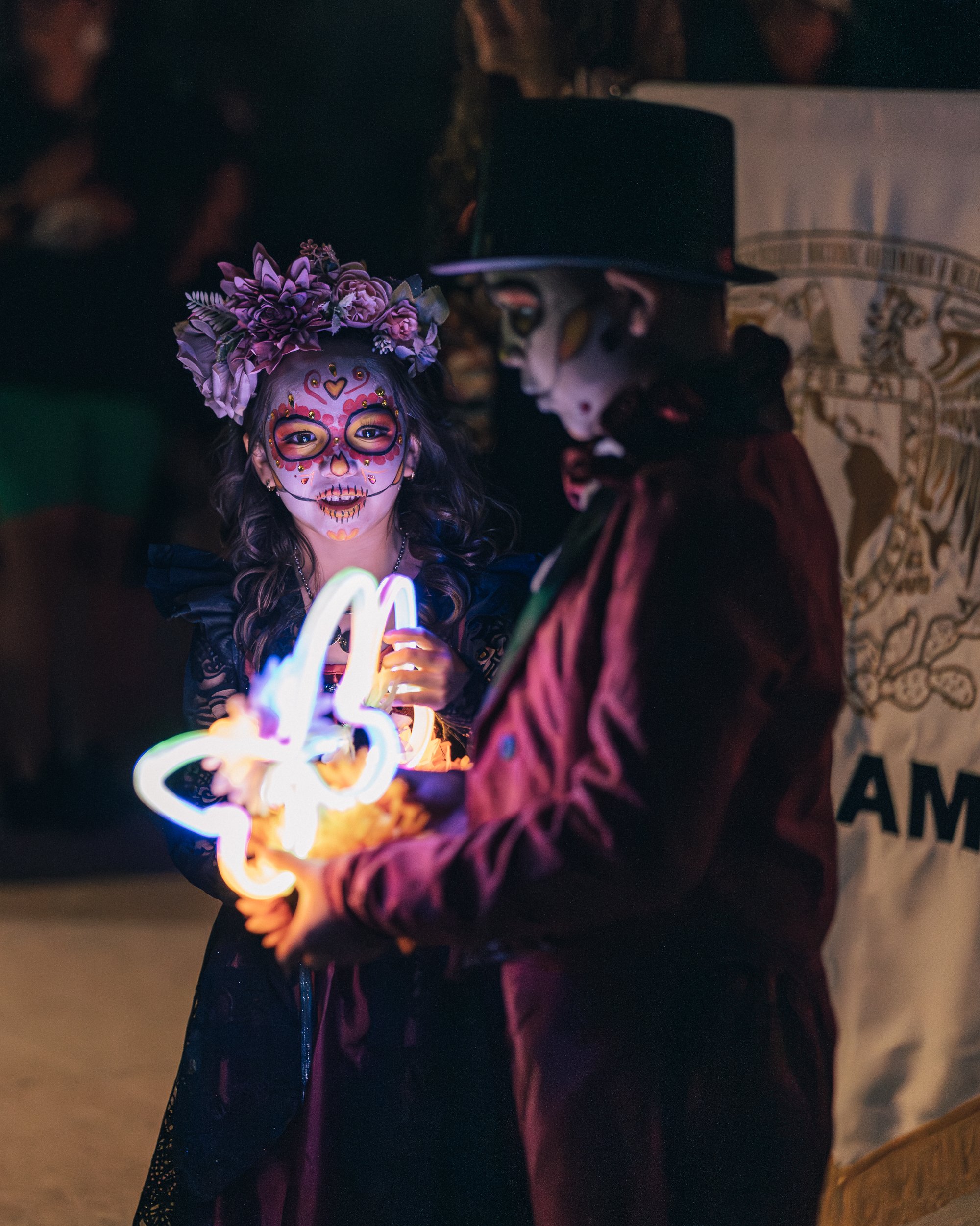 A young girl in Día de los Muertos face paint and a flower crown holds glowing butterfly-shaped lights while looking toward a costumed boy beside her.