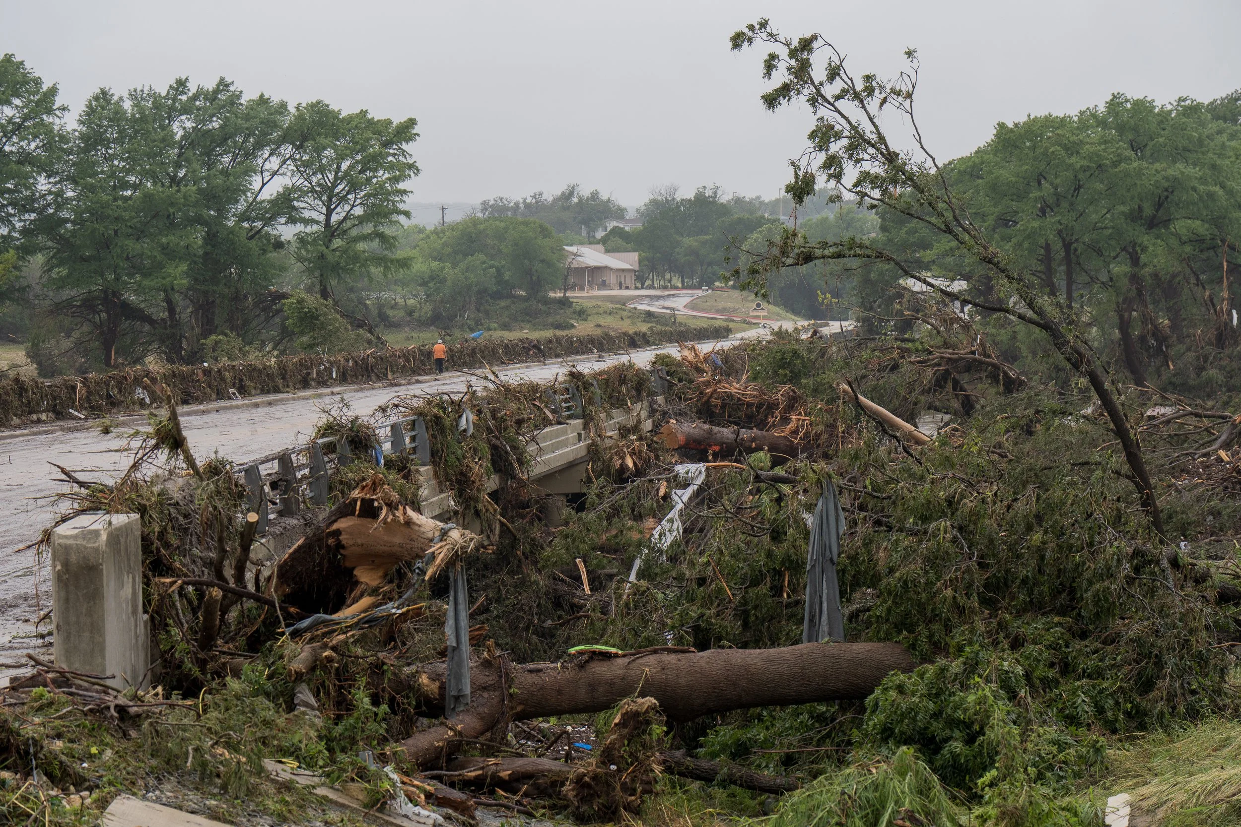 Debris and uprooted trees jam a damaged bridge after the Central Texas flash floods, with a lone worker in an orange vest in the distance.