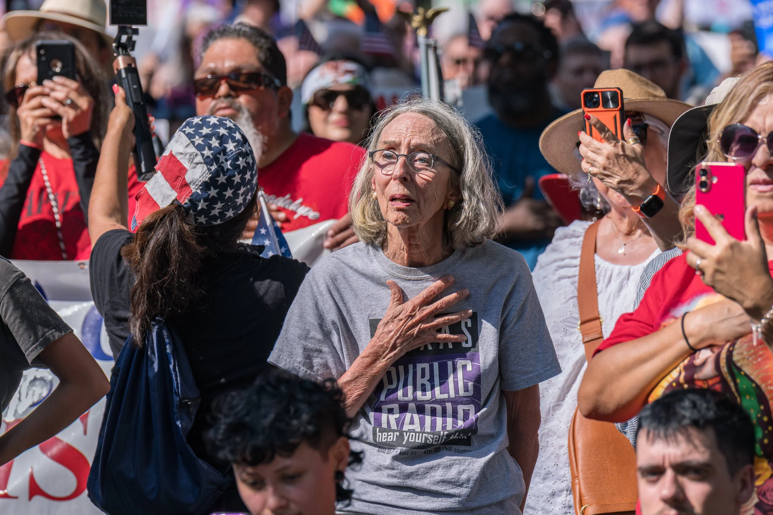 An older woman in a “Public Radio” T-shirt places her hand over her heart during the national anthem at the “No Kings” protest, surrounded by fellow demonstrators.