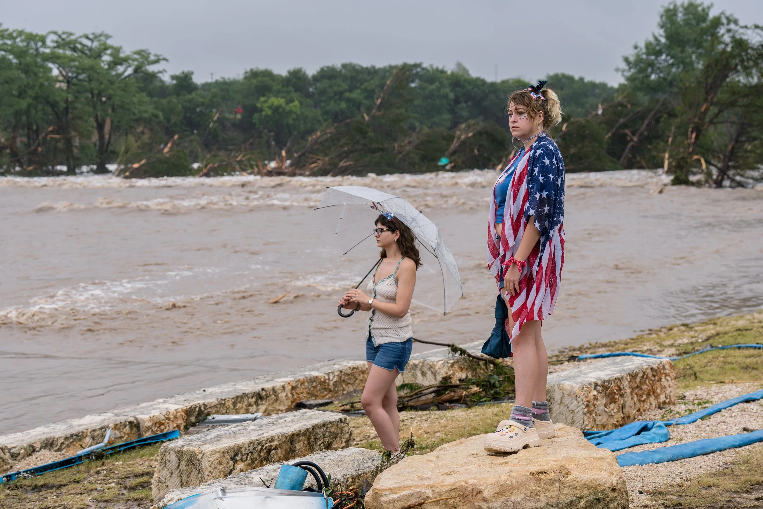 Two young women stand beside the flooded Guadalupe River, one wearing an American-flag poncho and the other holding a clear umbrella as they watch the high water in Kerrville, Texas.
