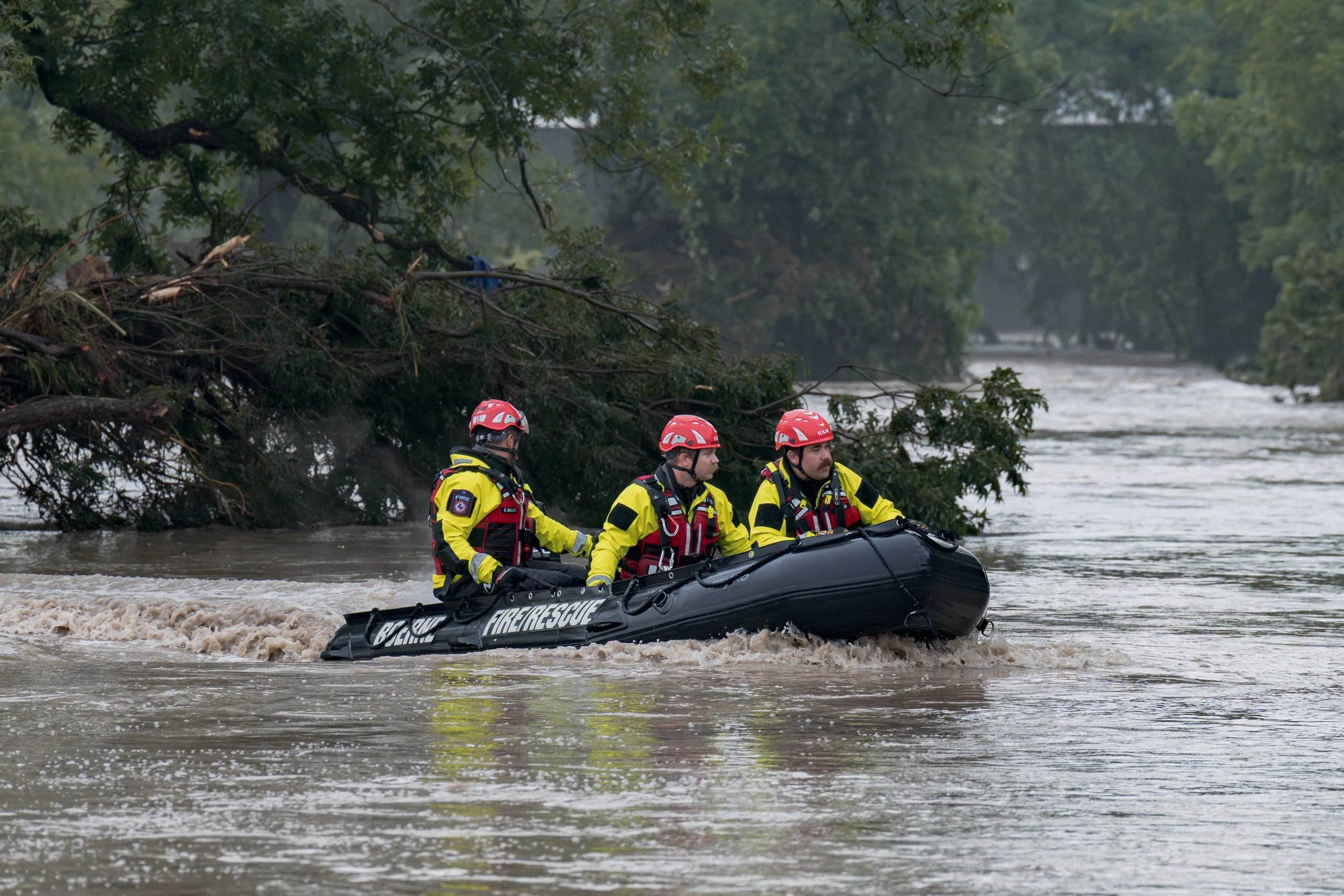 Boerne Fire Department swift-water rescuers navigate a black inflatable boat through fast-moving floodwater on the Guadalupe River lined with cypress trees.