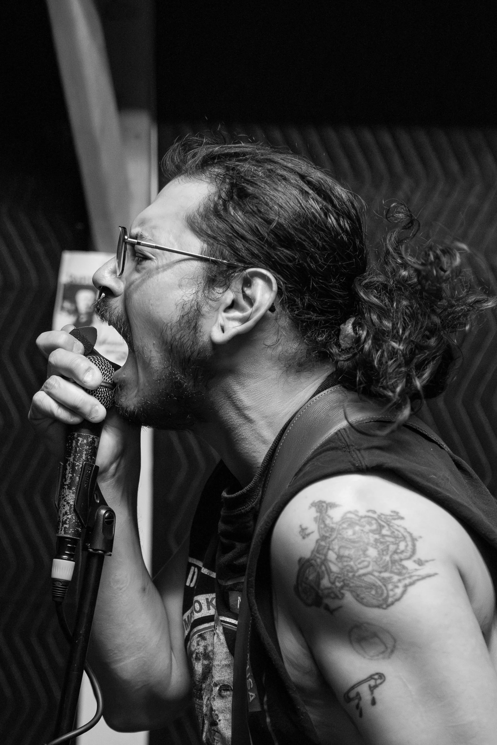 A punk vocalist screams into a handheld microphone, neck muscles taut, while performing in a small, dark rehearsal space.