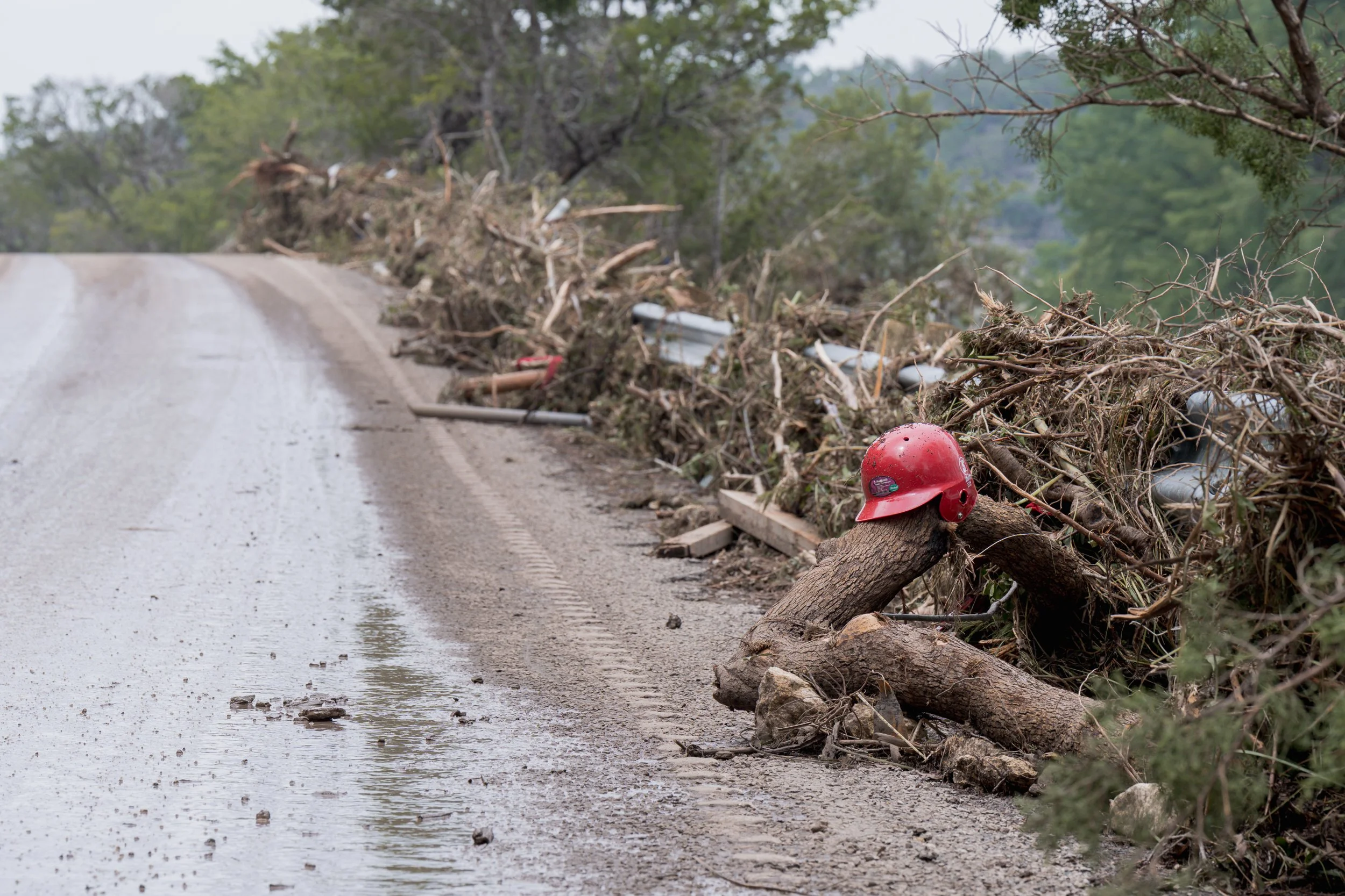A child's baseball helmet rests on a splintered tree trunk amid branches and mud piled beside a rural road after the Central Texas flash floods.