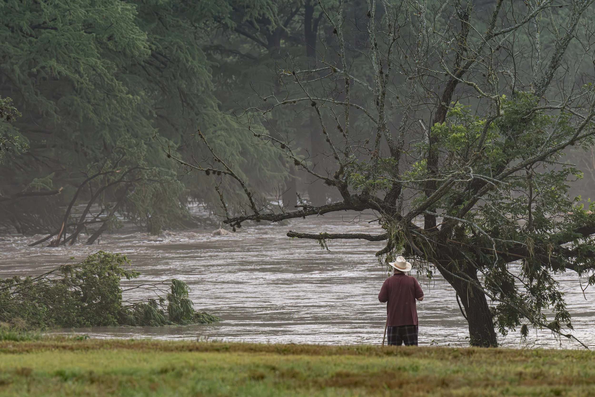 A Kerrville resident in a tan hat and maroon jacket stands beside the swollen Guadalupe River, watching floodwaters rise during the Central Texas floods.