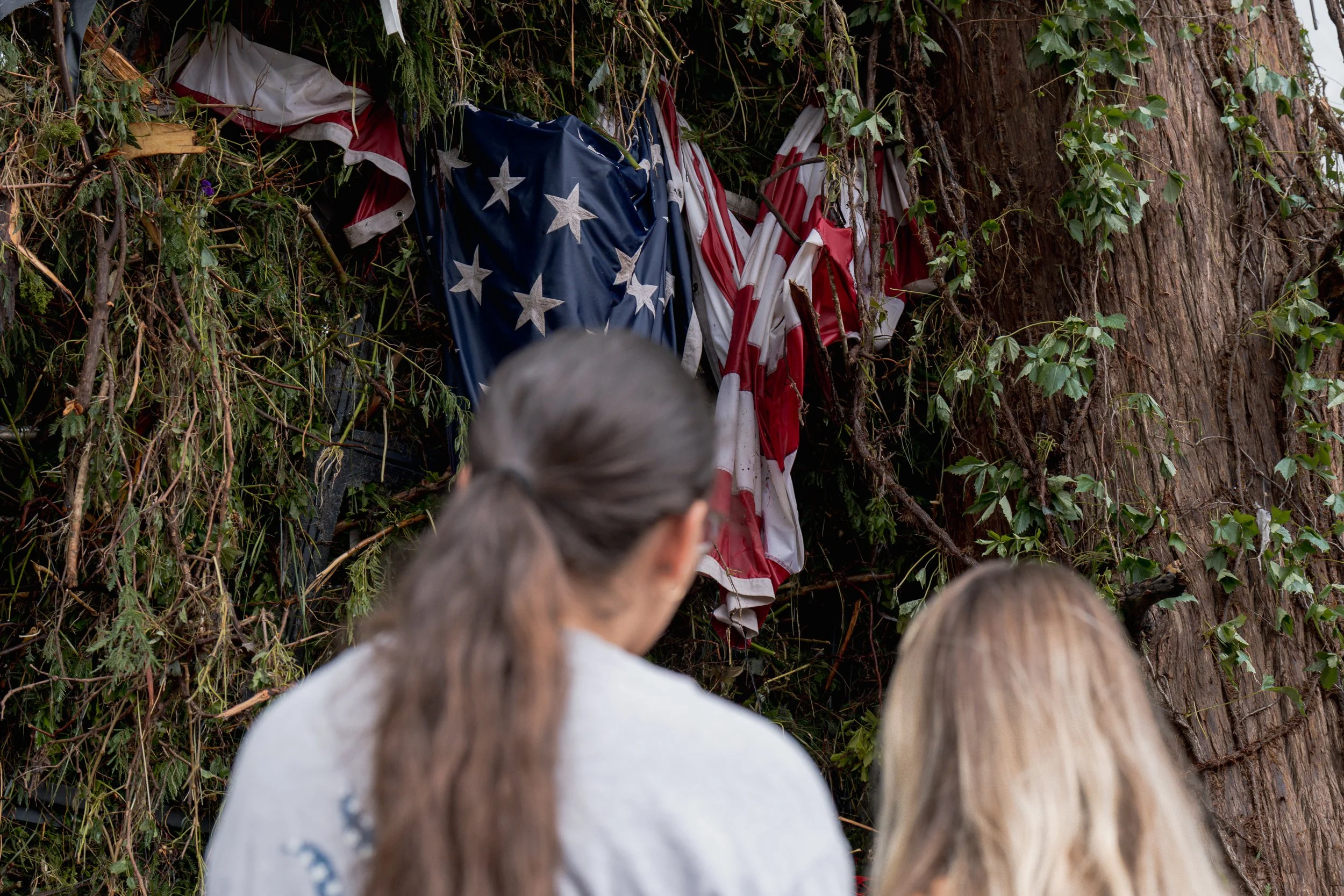 Two people stand facing a large tree draped with torn, weathered American flags left hanging after the Central Texas flooding.