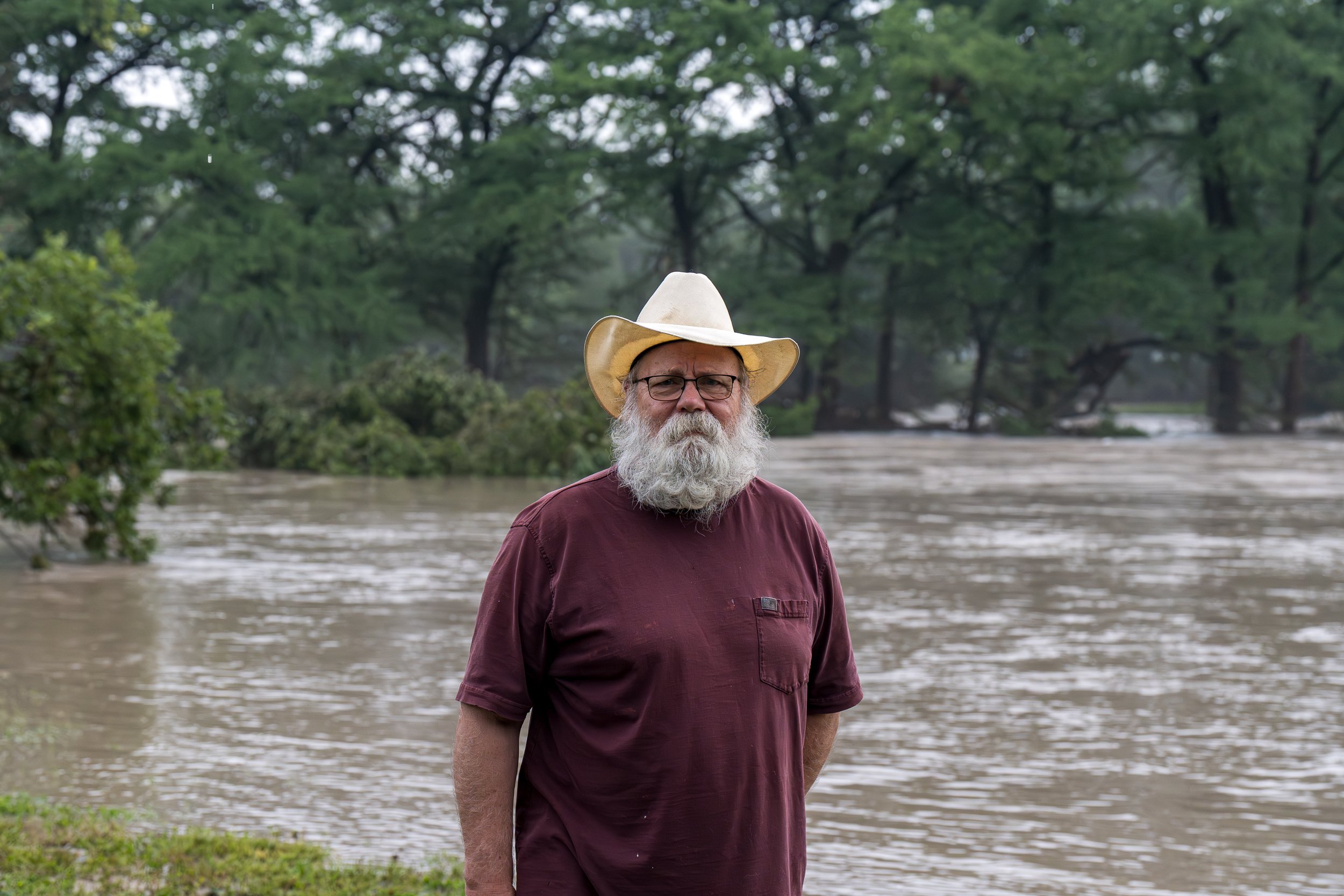 A bearded man in a maroon shirt and white cowboy hat stands in front of the swollen Guadalupe River on a rainy day in Central Texas.