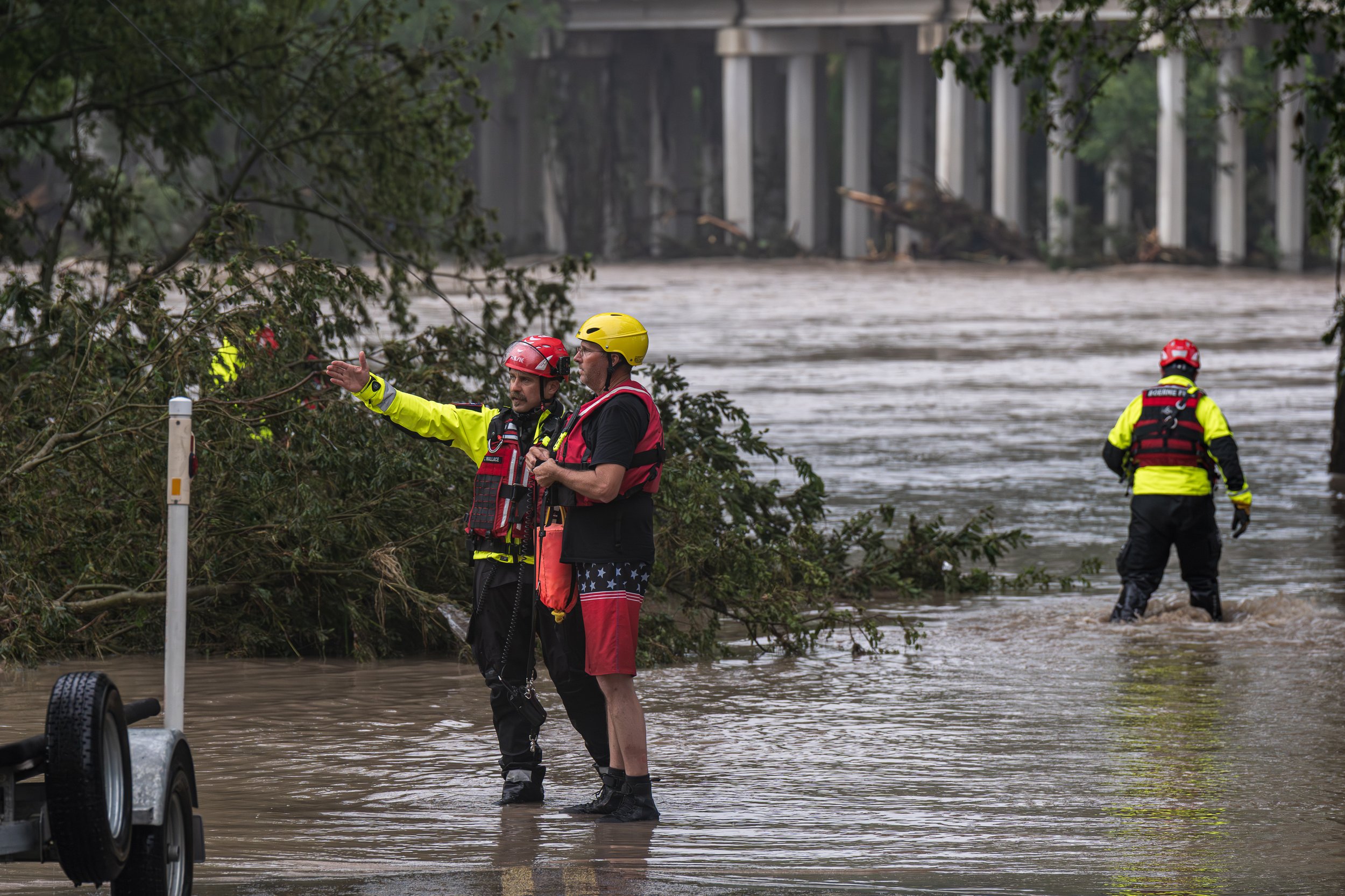 Rescue workers in helmets stand in floodwater near downed trees as a person on the road points toward the flooded area.