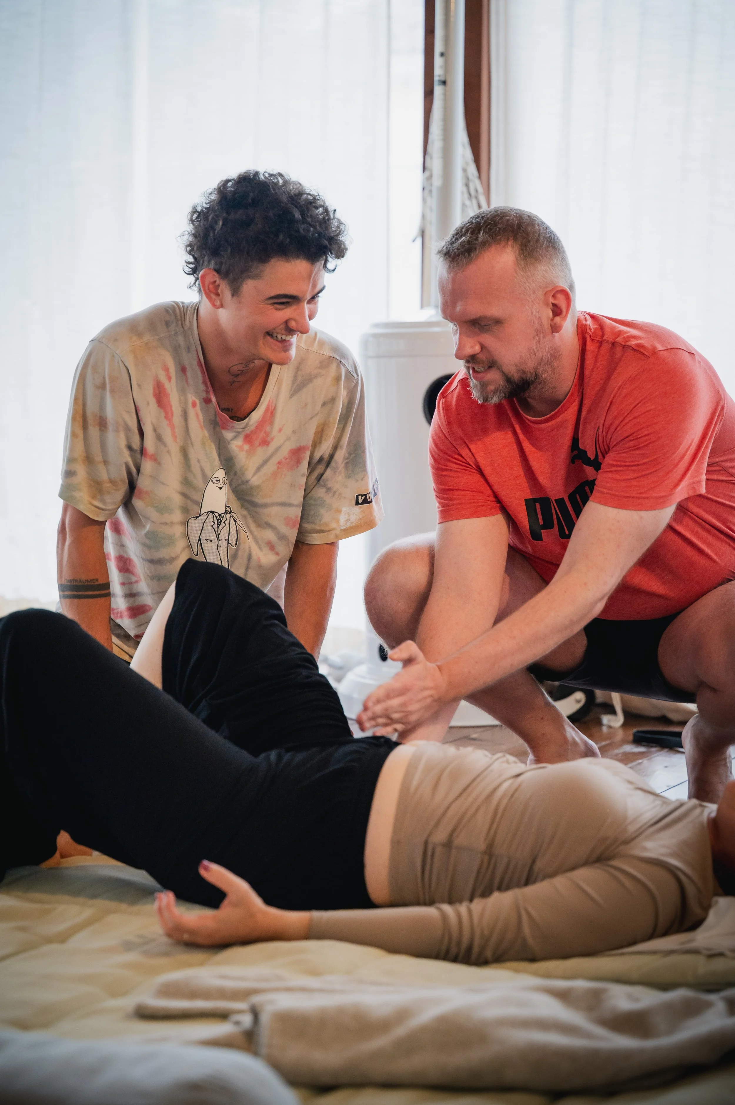 Two young men are playing with a woman lying on a bed. The men are smiling and appear to be having fun.